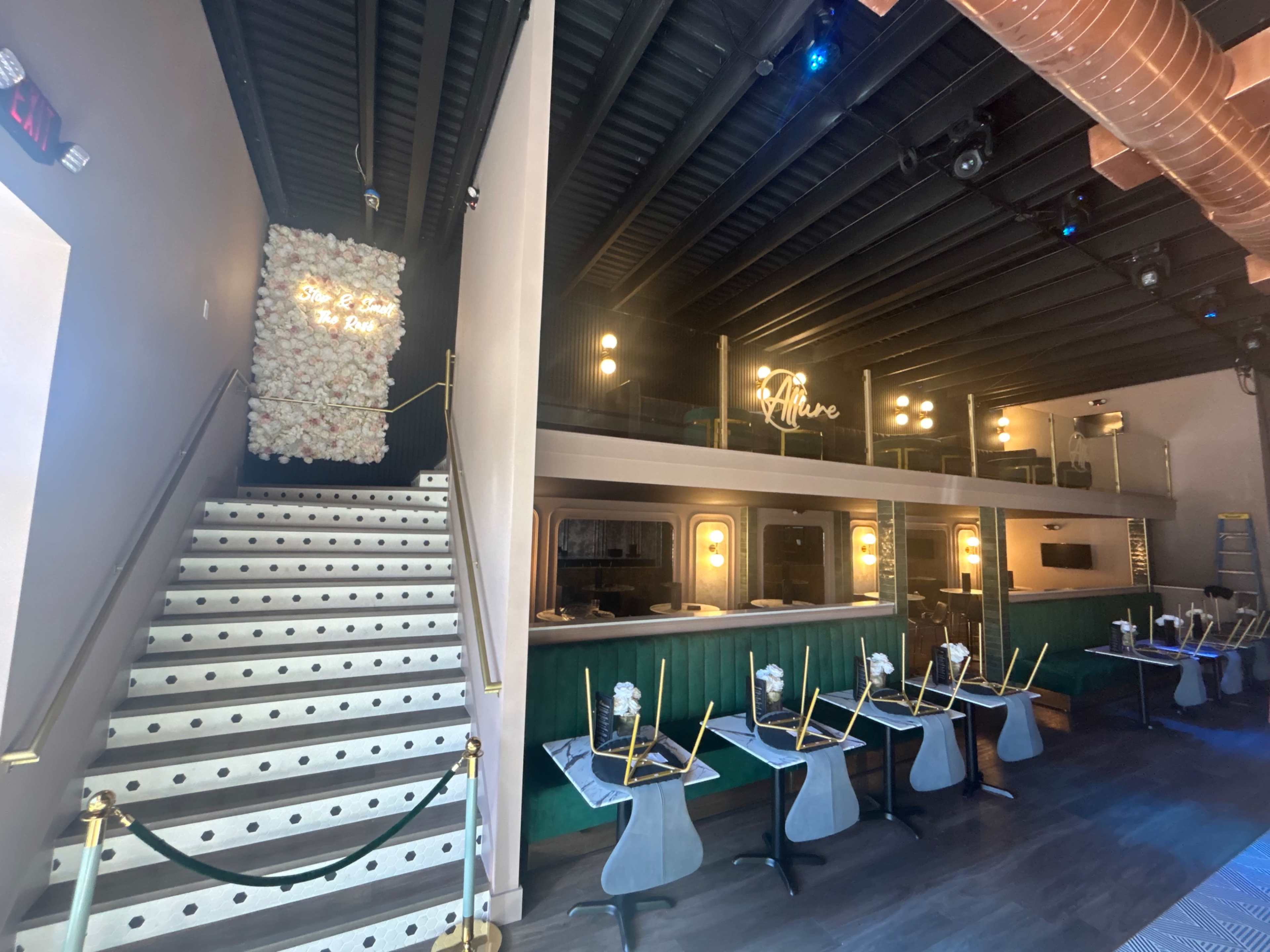 The image shows a modern restaurant interior featuring a staircase, a bar area, and decorative floral arrangements on the wall.