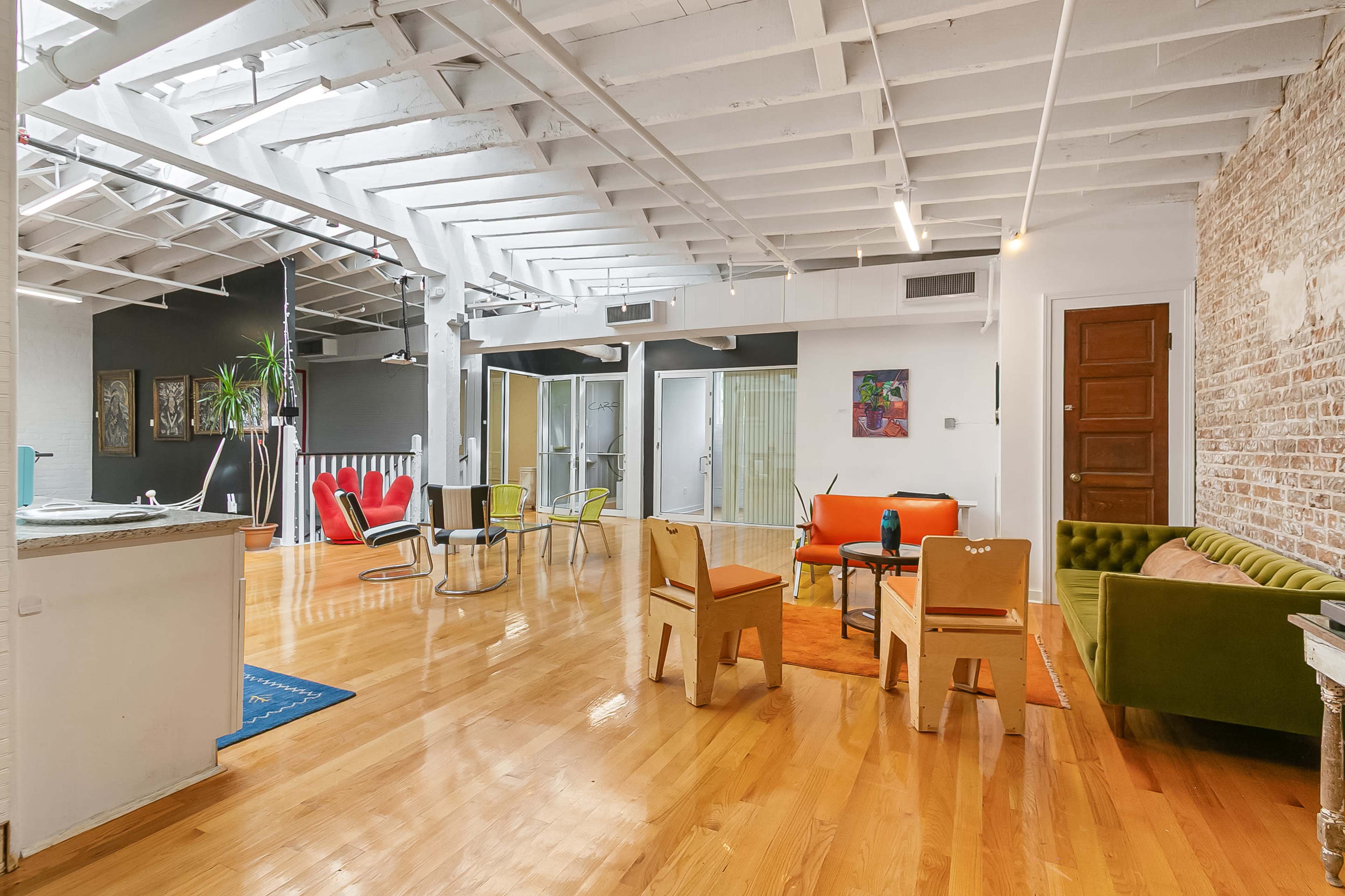 The image shows a spacious, open-plan living area with hardwood floors, a mix of colorful furniture, and exposed brick walls.