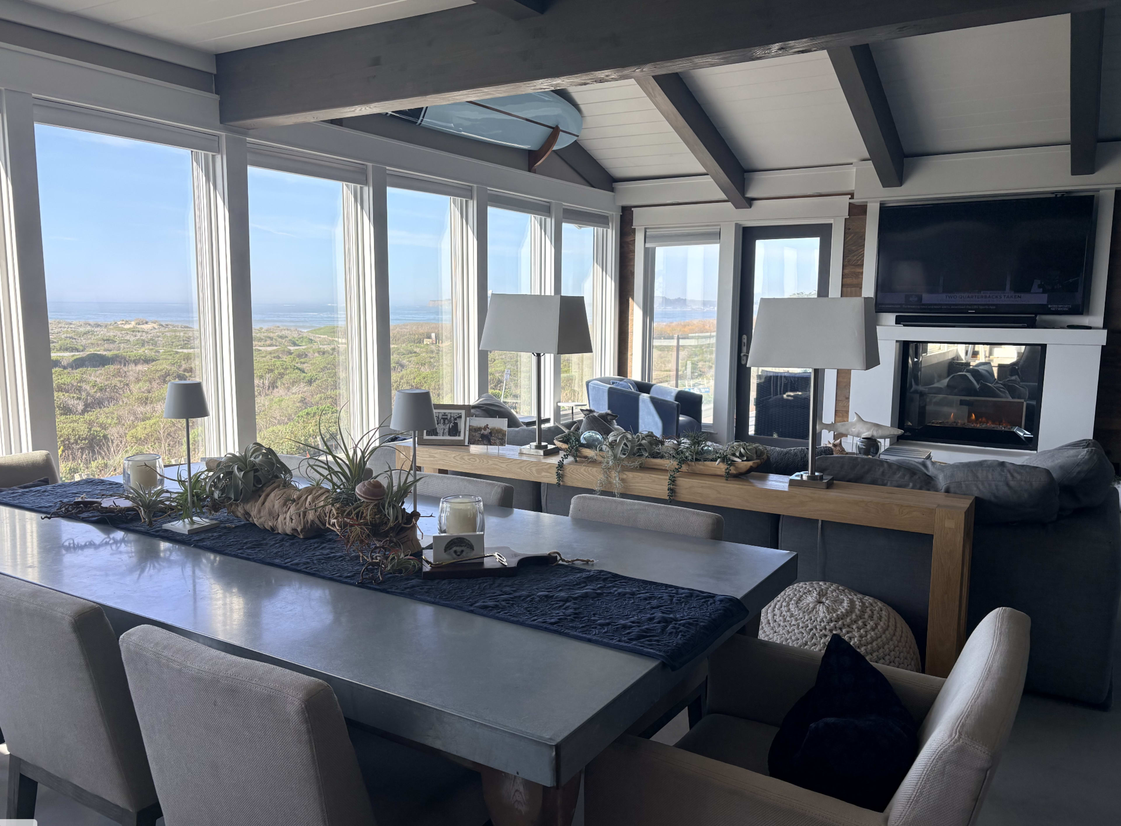 The image shows a modern, open-concept living space with large windows overlooking a landscape, featuring a dining table, seating area, and a fireplace.