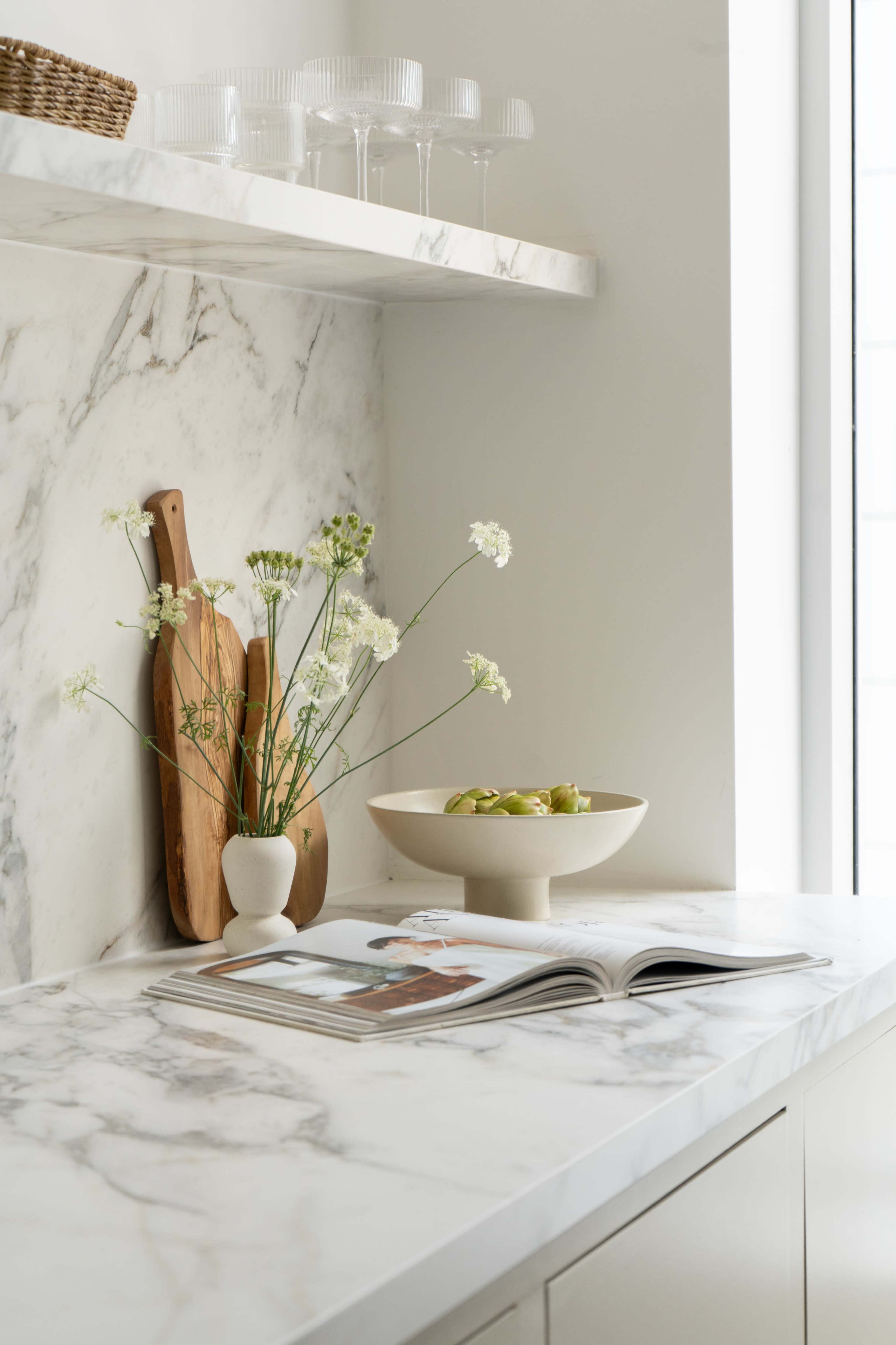 A modern kitchen countertop features a marble surface, wooden cutting boards, a vase with flowers, and a bowl of fruit, alongside an open magazine.