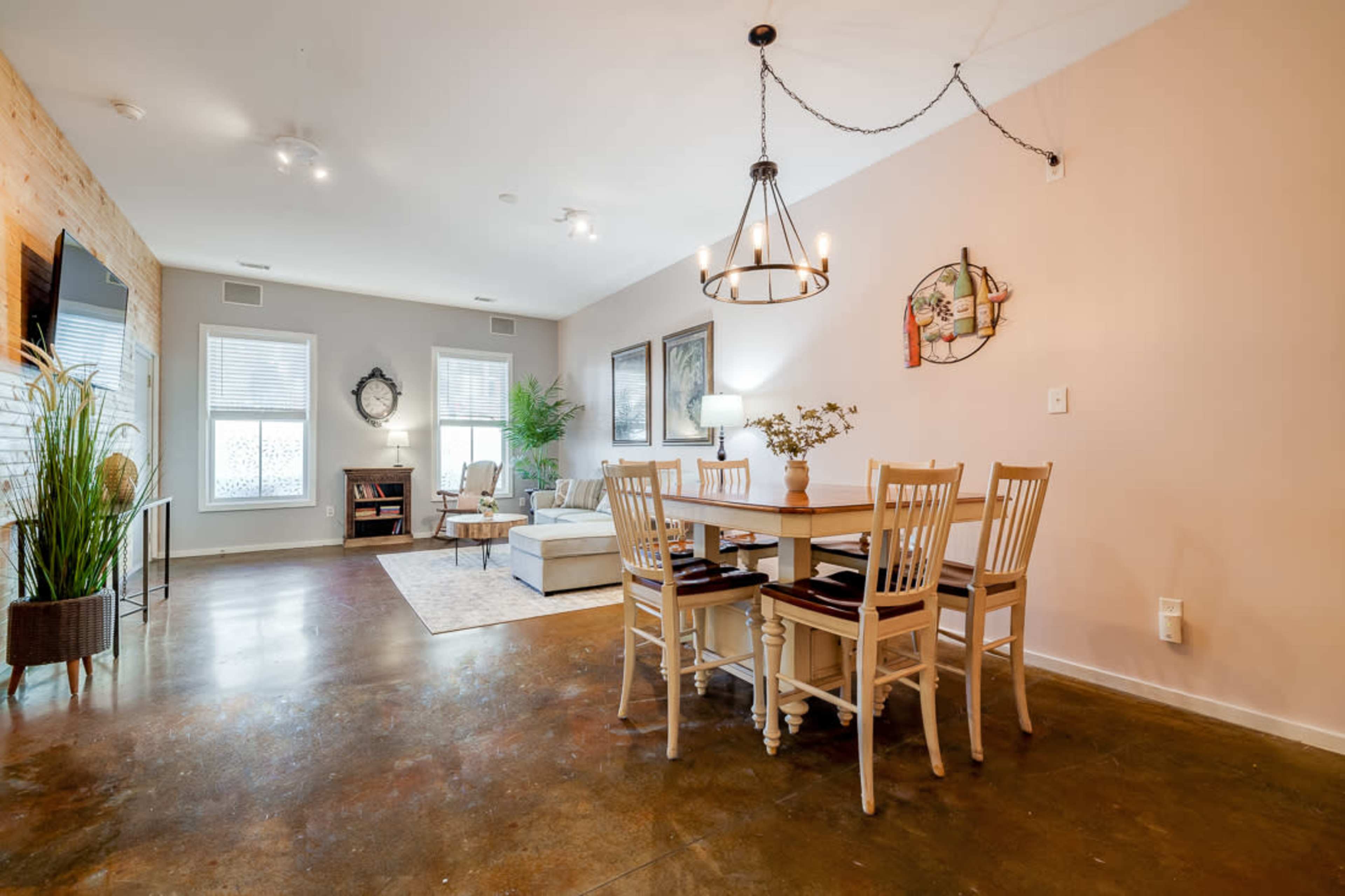 A spacious living and dining area features a table with six chairs, a sofa, and decorative plants against neutral-colored walls.