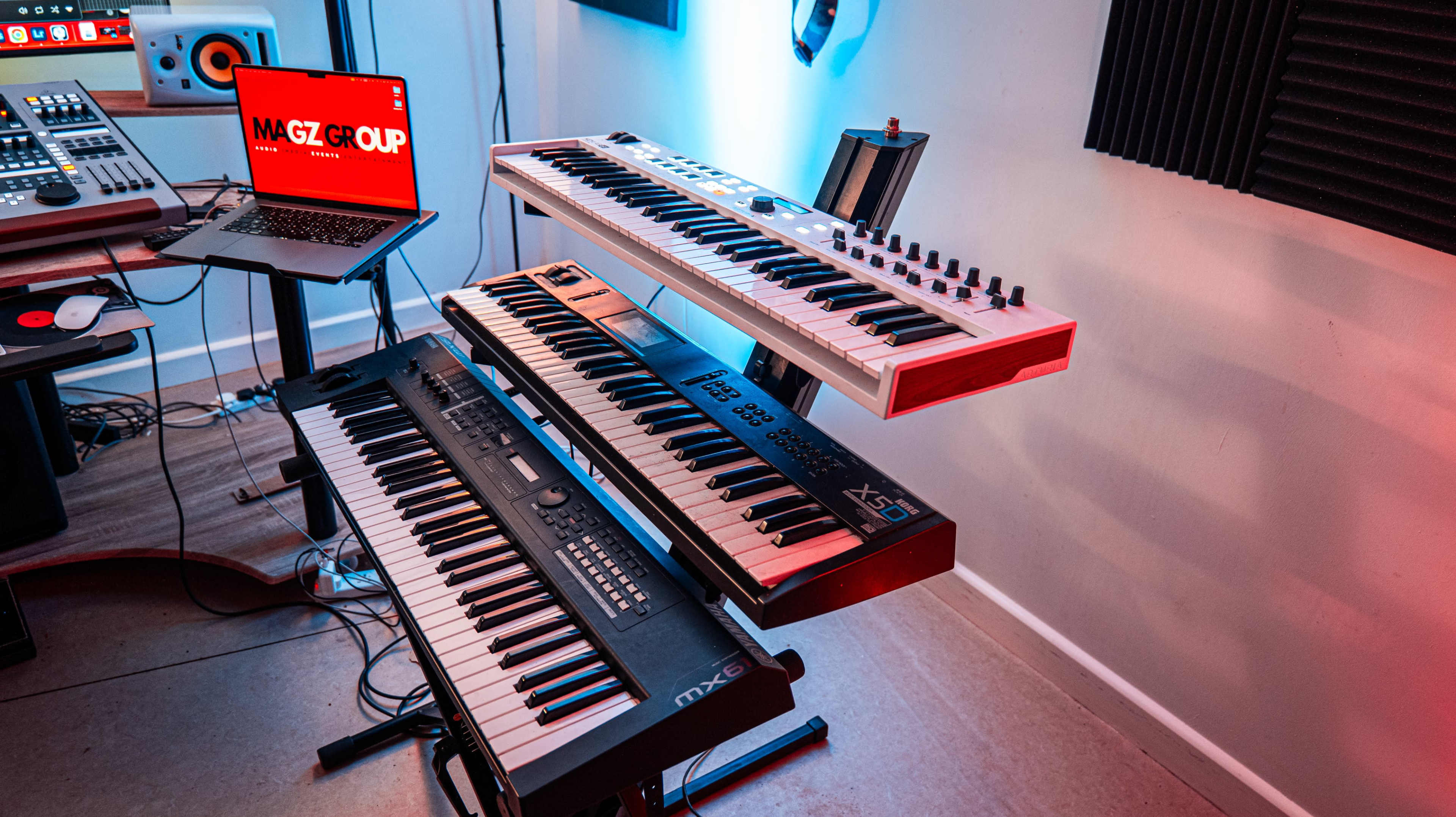 The image shows a music production setup featuring three keyboards arranged on a stand, with a laptop positioned nearby.