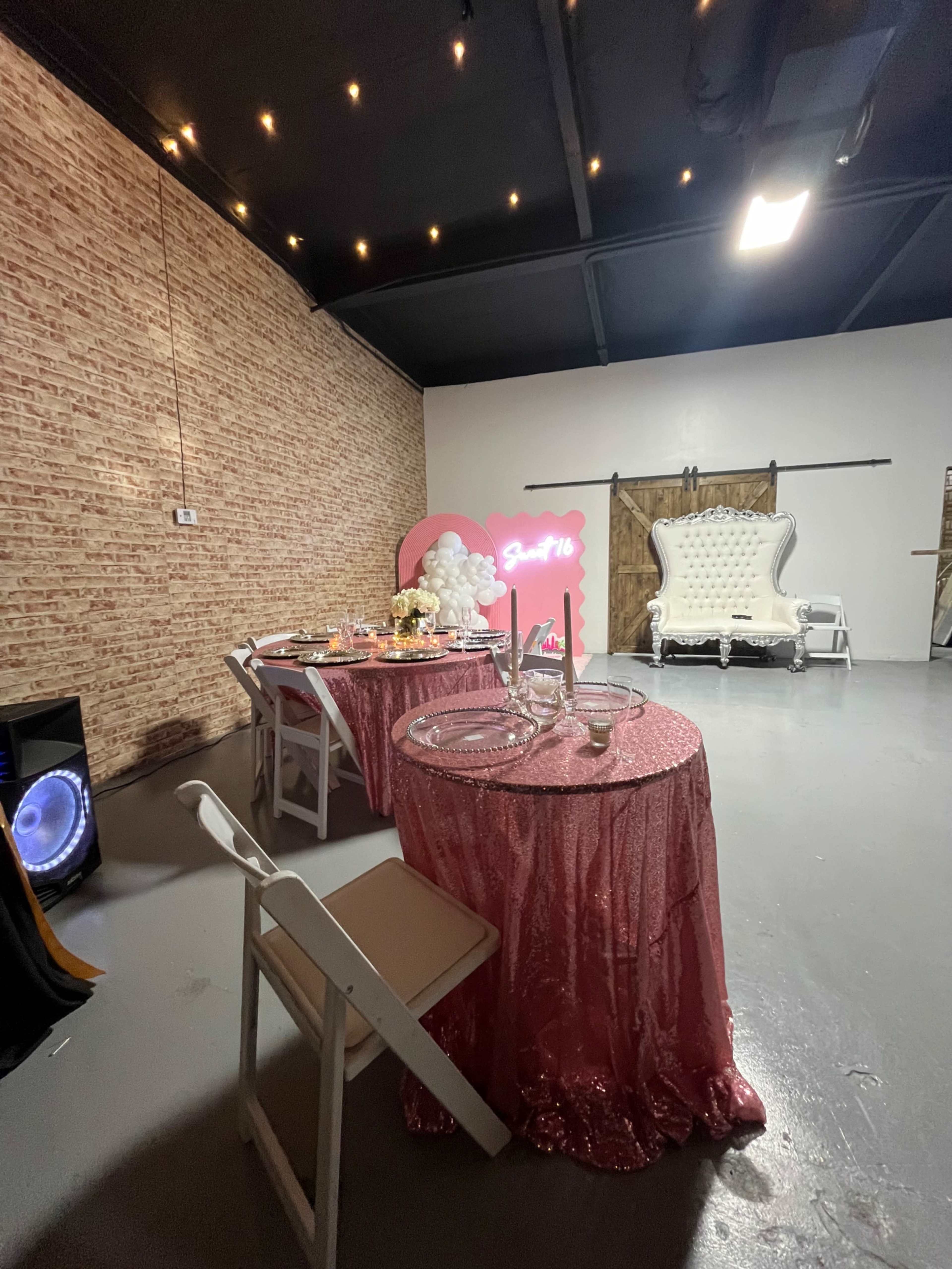 The image shows a decorated event space with a pink-themed table setup, a plush white couch, and a neon sign against a brick wall.