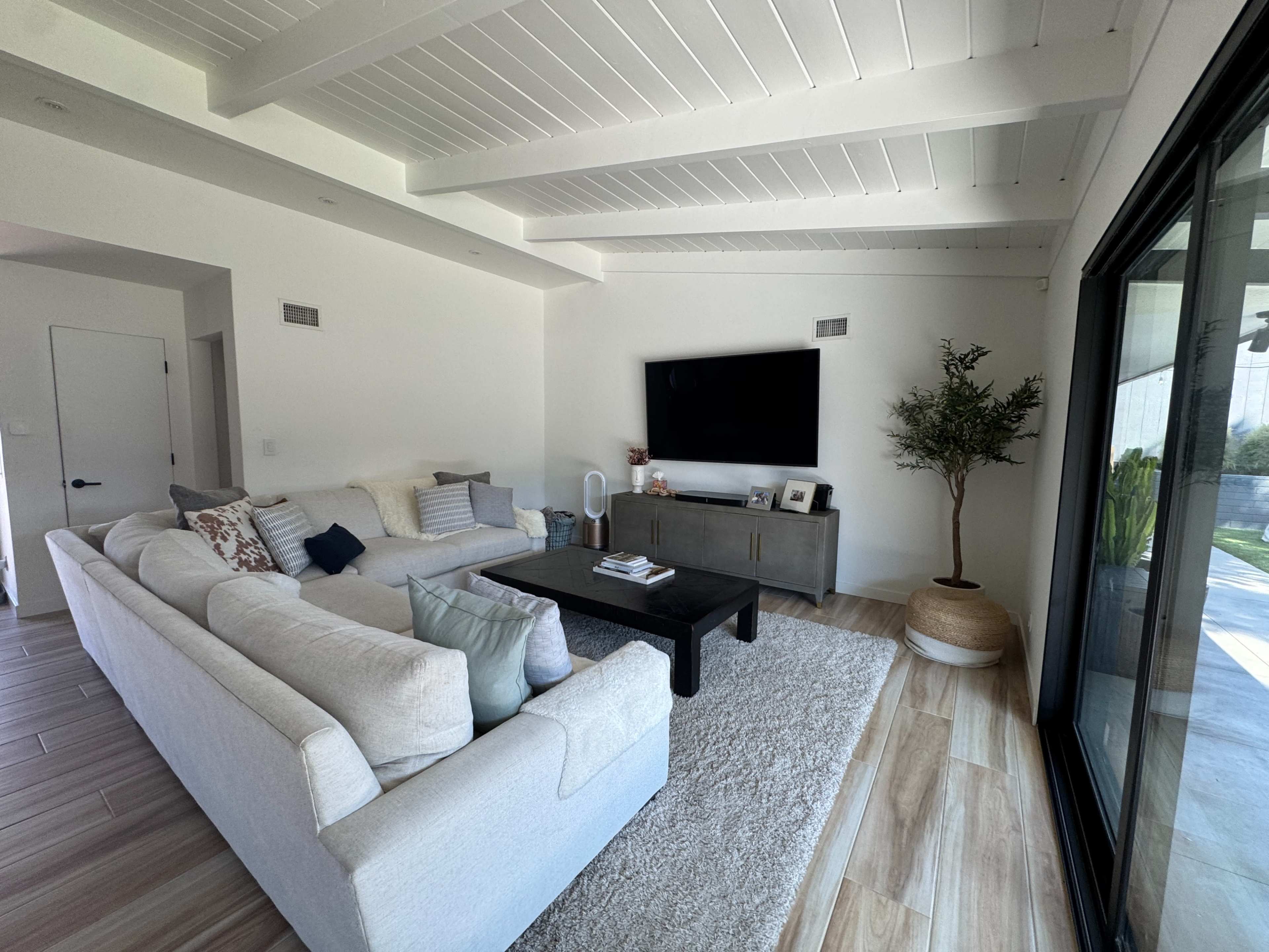 A modern living room features a large sofa, a dark coffee table, a television mounted on the wall, and a potted plant in the corner.
