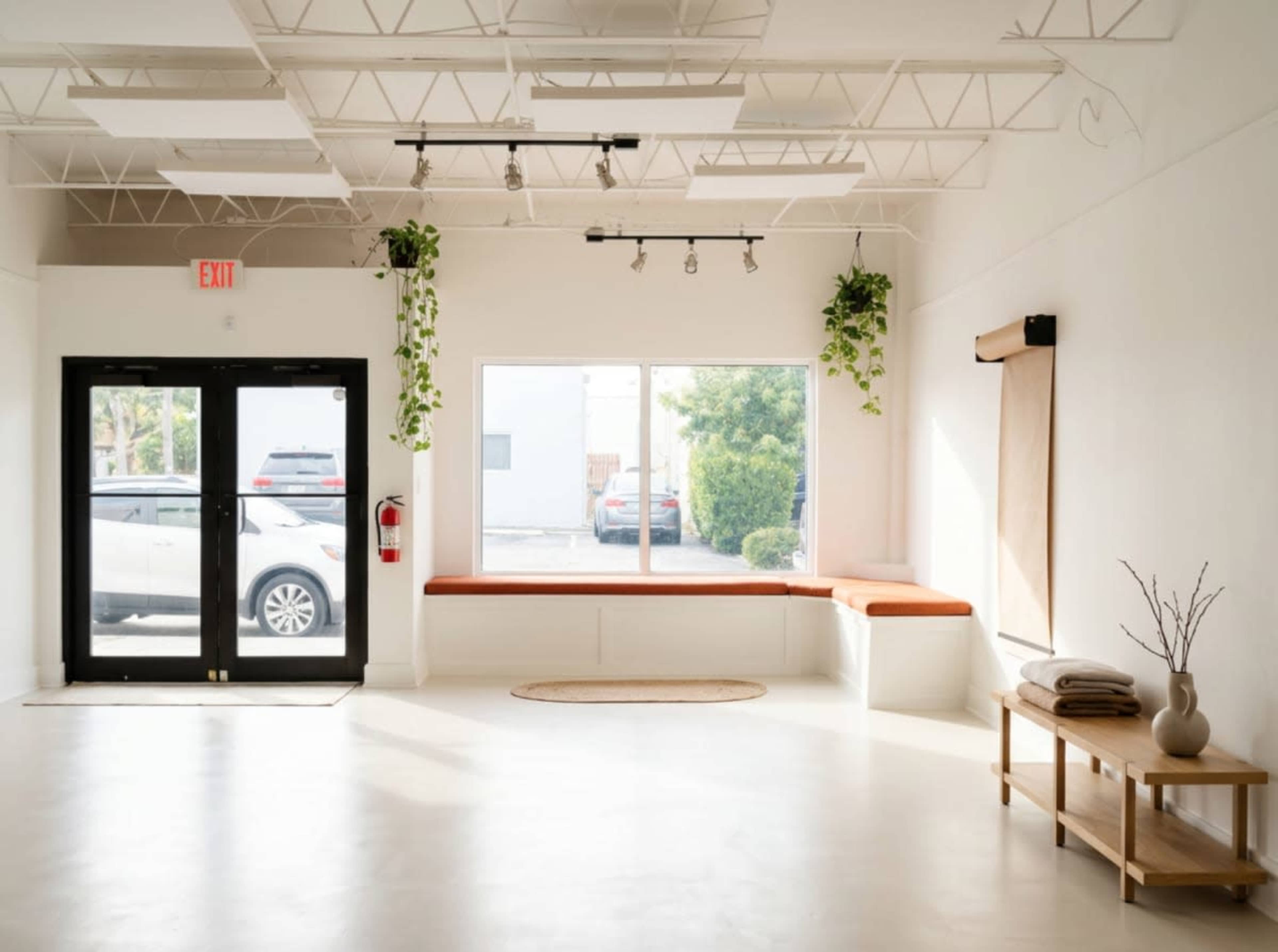 The image shows a spacious, minimalist interior of a room with a large window, a bench with orange cushions, and a wooden table decorated with a vase.