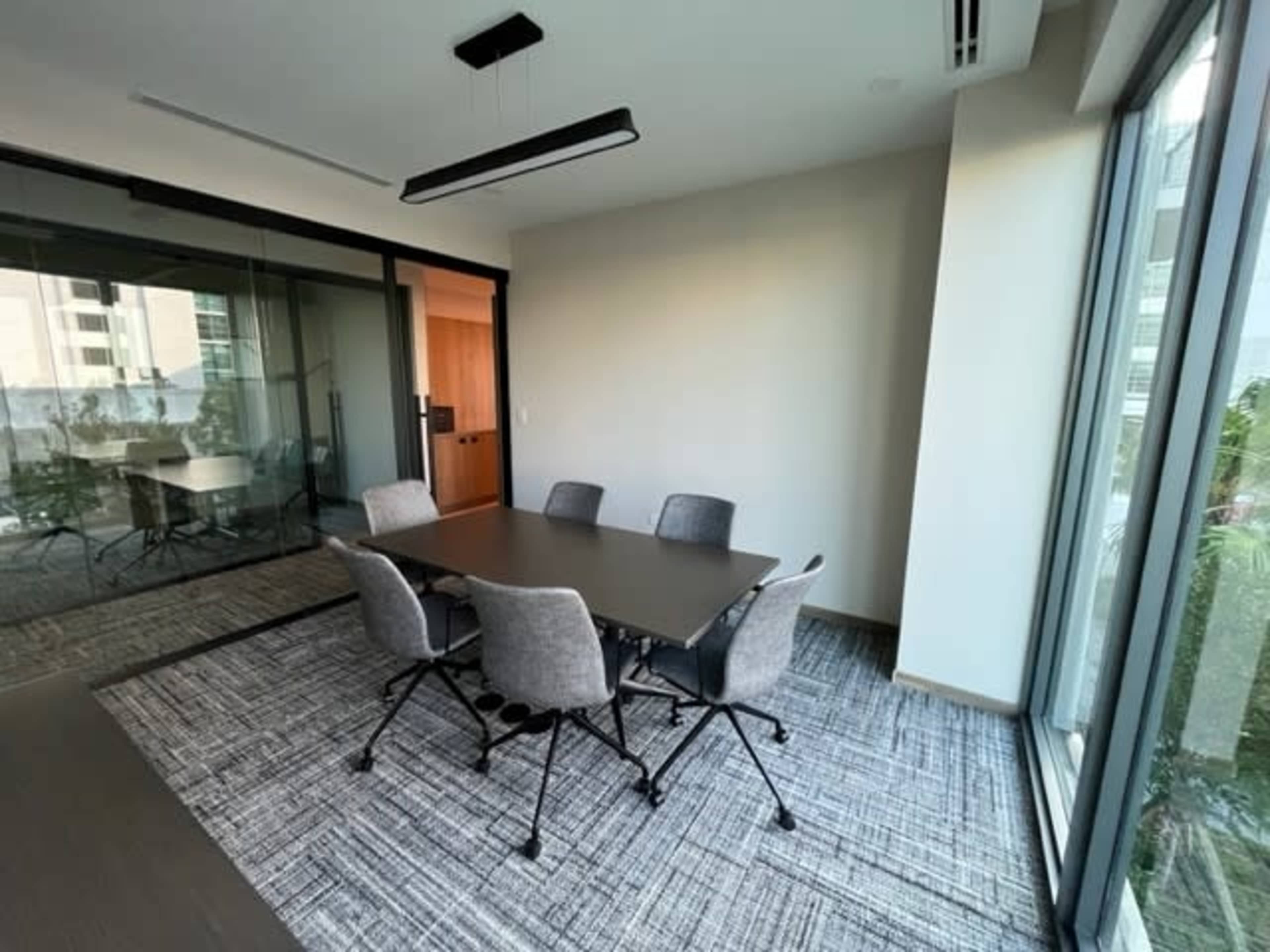 A conference room features a rectangular table surrounded by six chairs, with large windows offering a view of the outside.