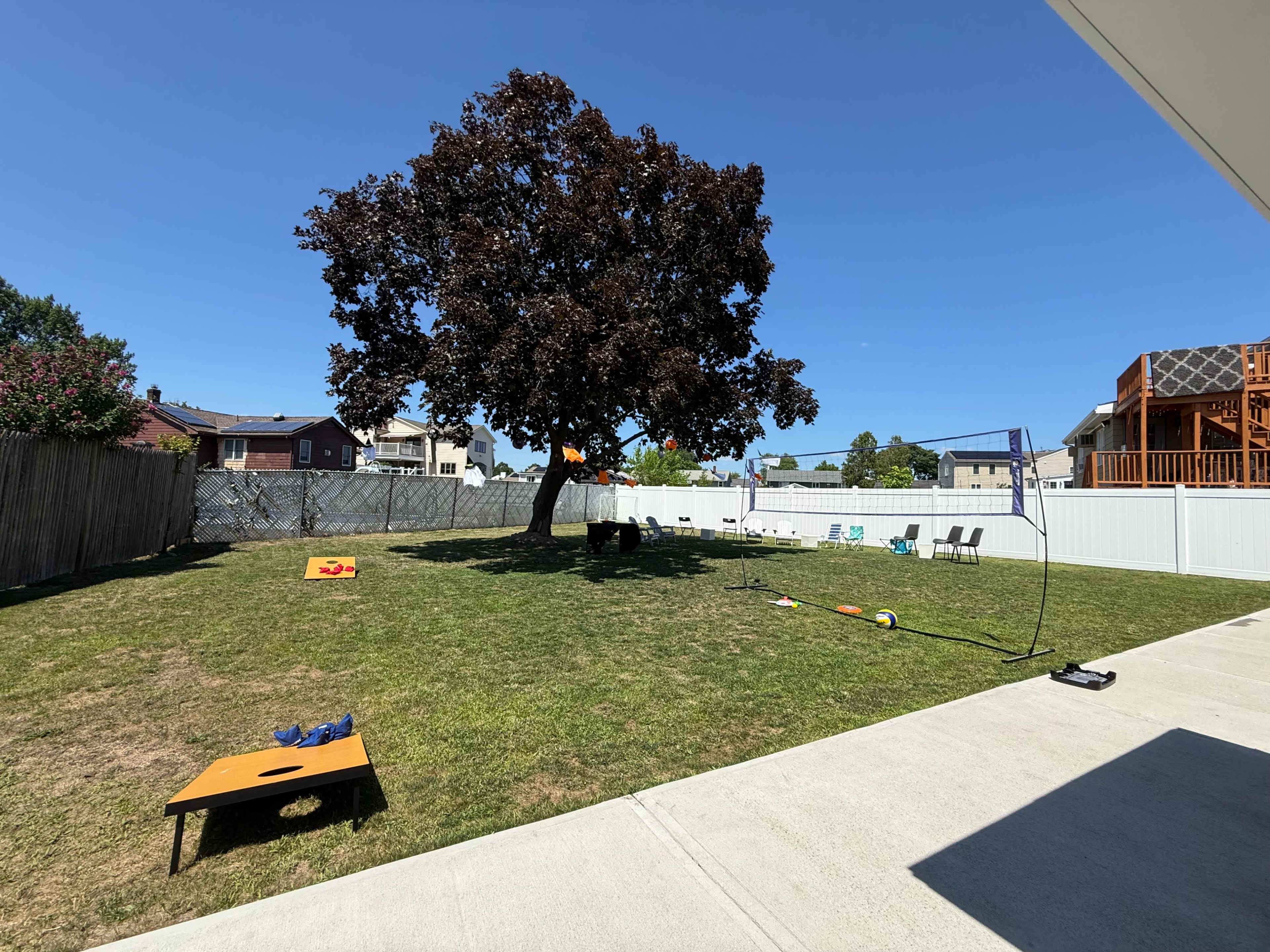 A large tree stands in a grassy yard with various outdoor games set up, and a residential area in the background.