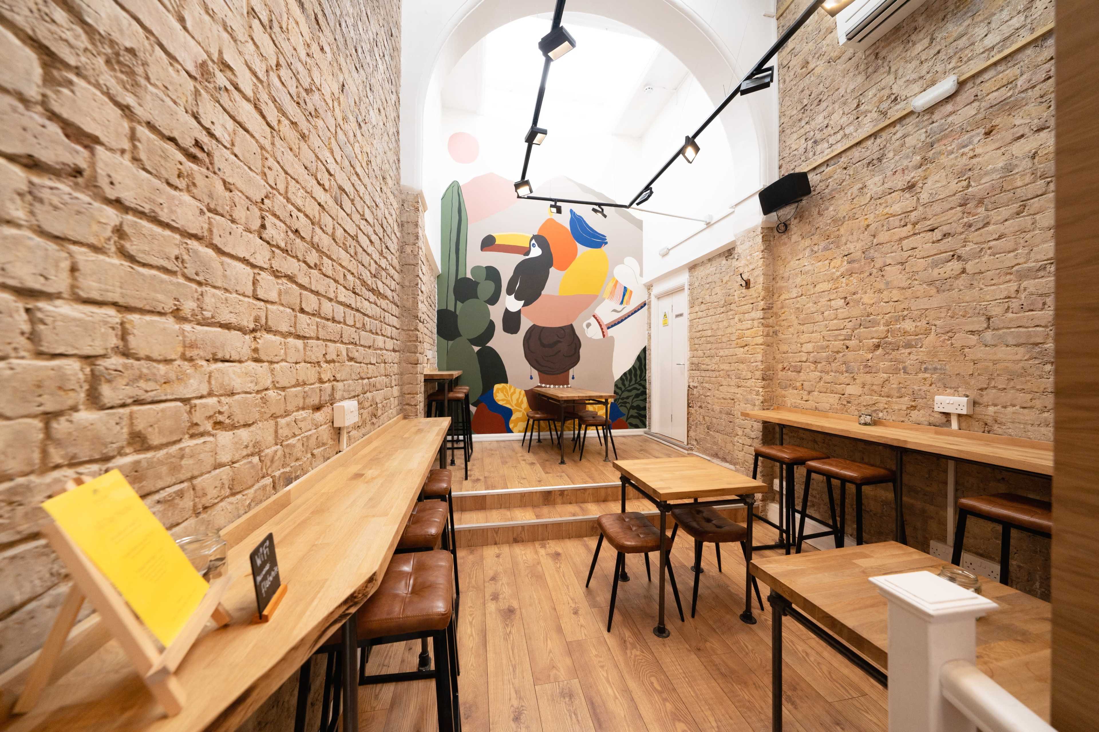 A narrow, well-lit restaurant with wooden tables and chairs, featuring a colorful mural on the back wall.