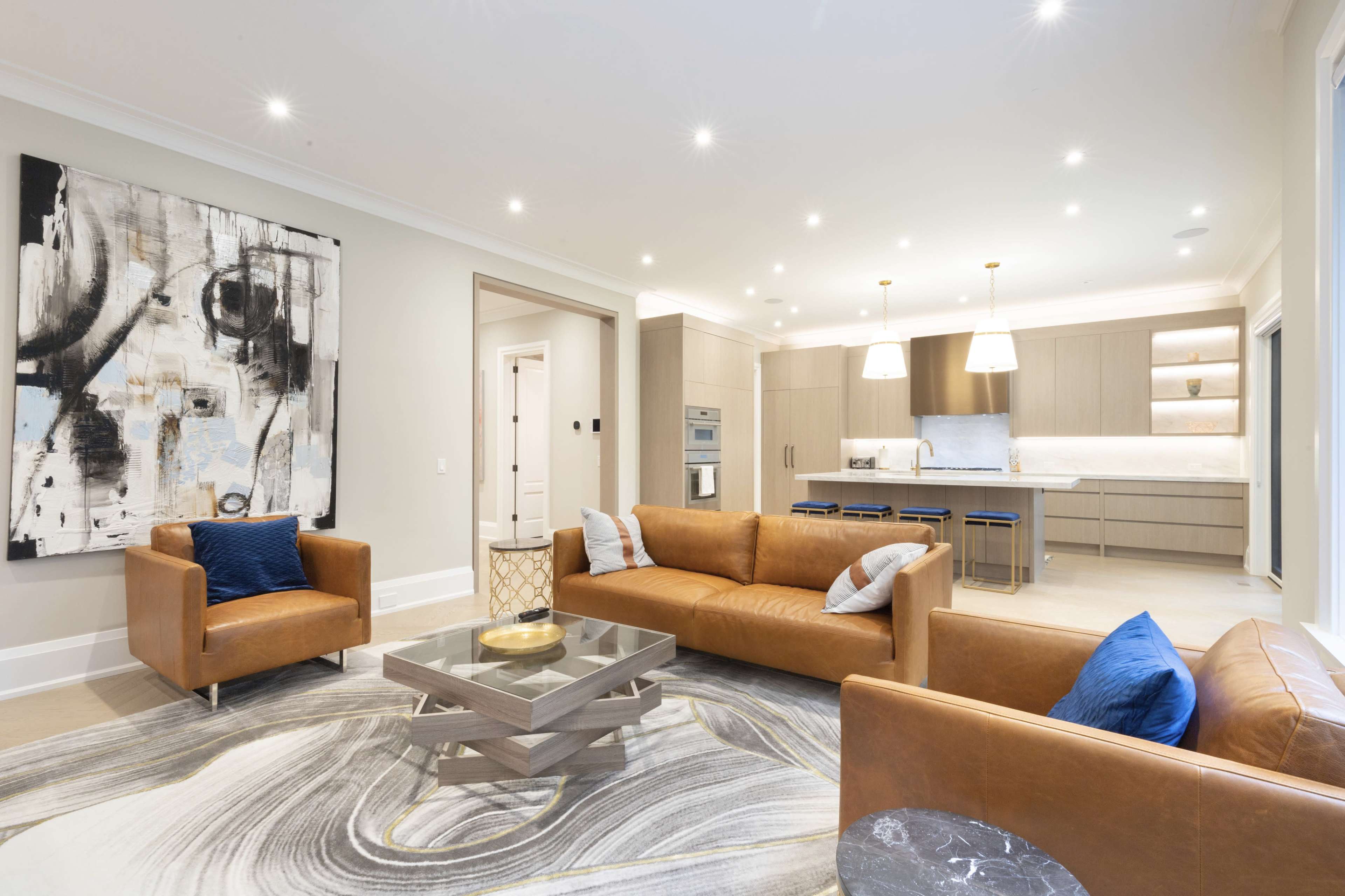 A modern living area with two leather sofas, a glass coffee table, and a large abstract painting on the wall, leading into an open kitchen with light-colored cabinetry.