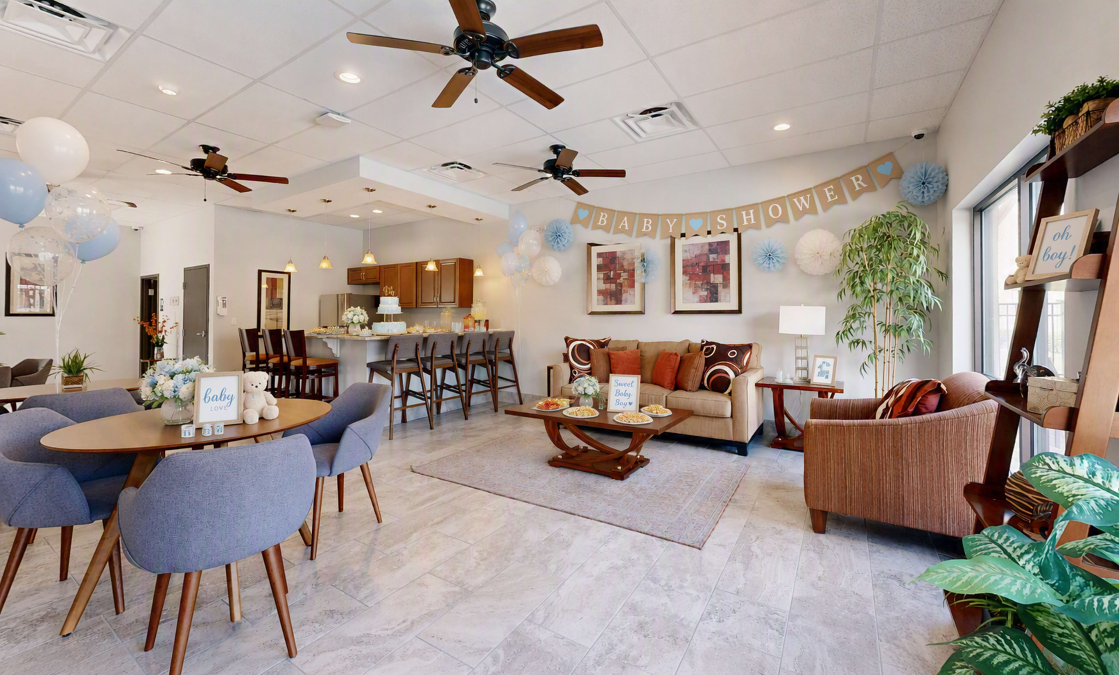 A decorated event space features a seating area with a couch and chairs, tables set for dining, and a banner reading "Baby Shower."