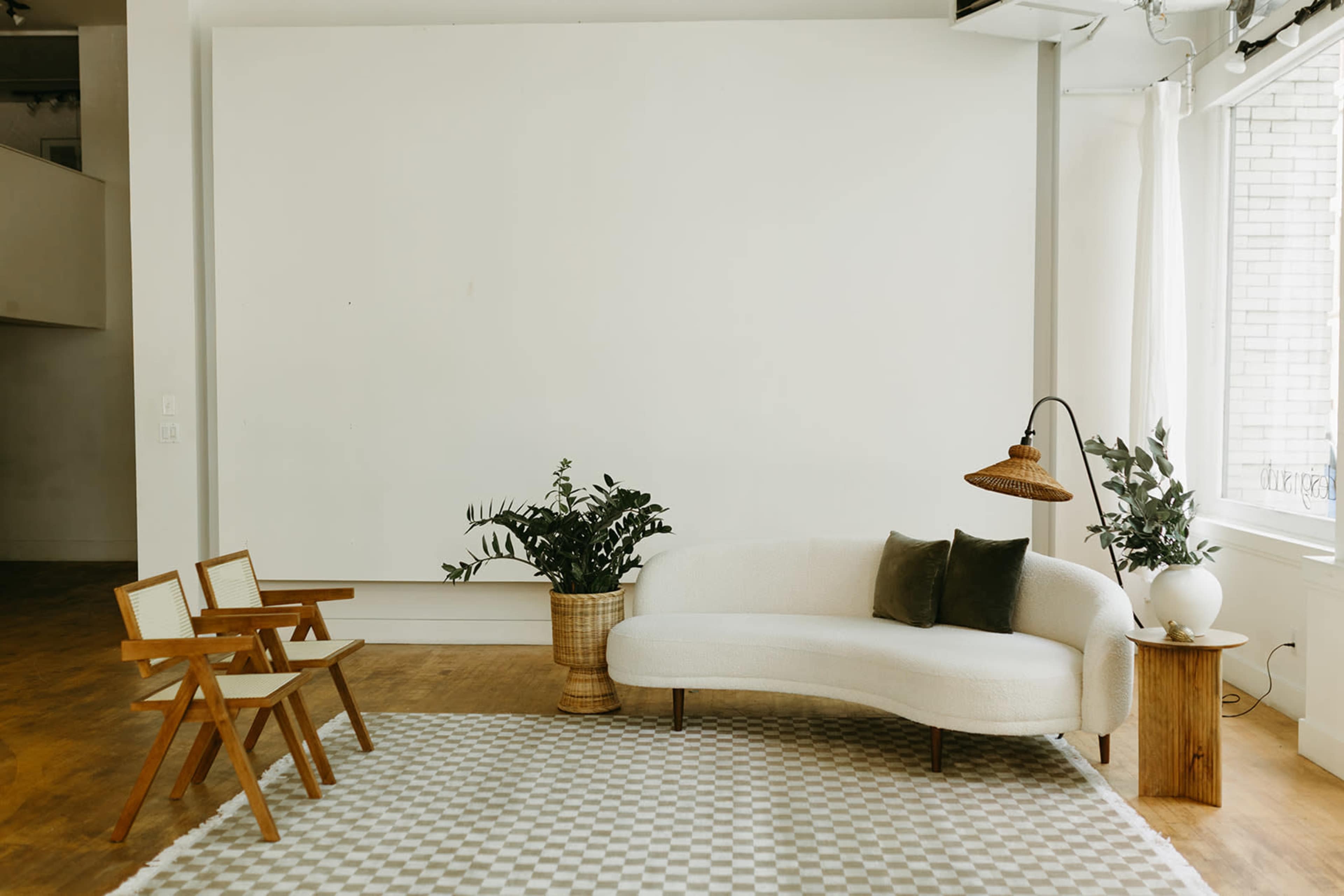 The image features a minimalist interior with a white wall, a light-colored curved sofa, two wooden chairs, a potted plant, and a woven lampshade.