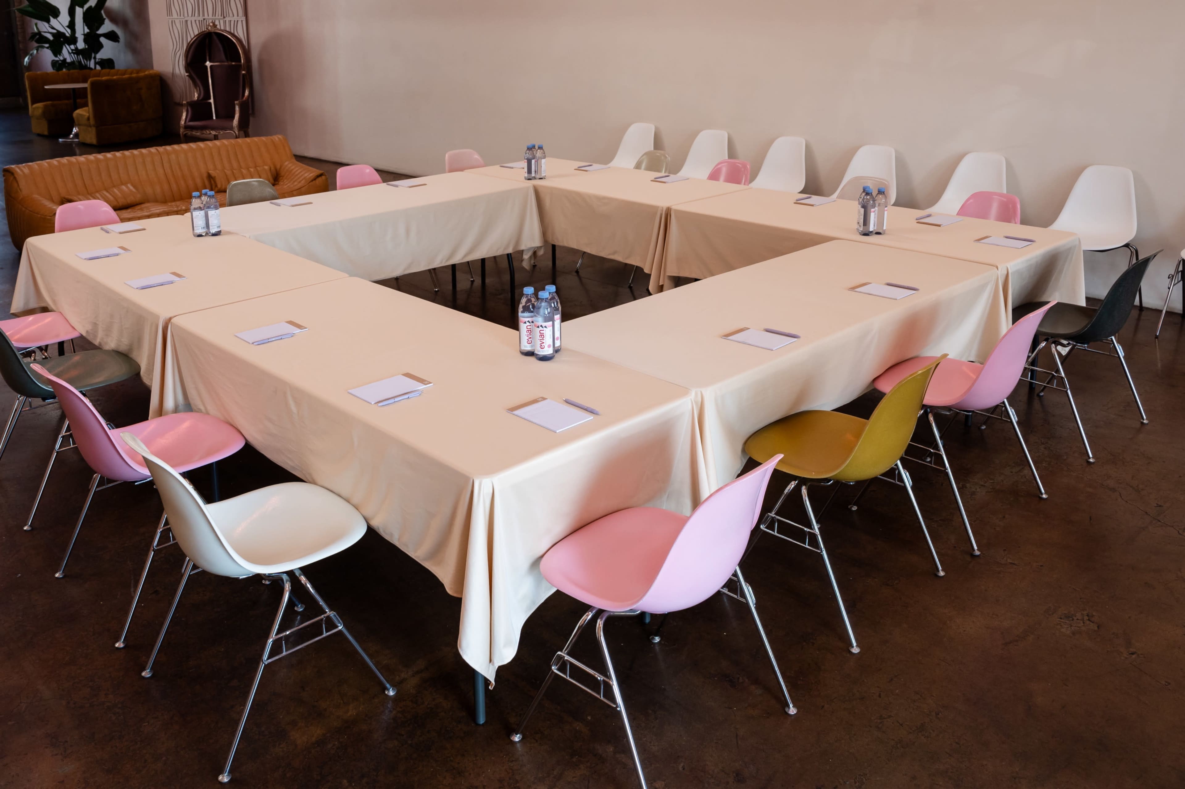 A rectangular meeting setup with light-colored tablecloths and chairs in various pastel colors surrounds a central area.