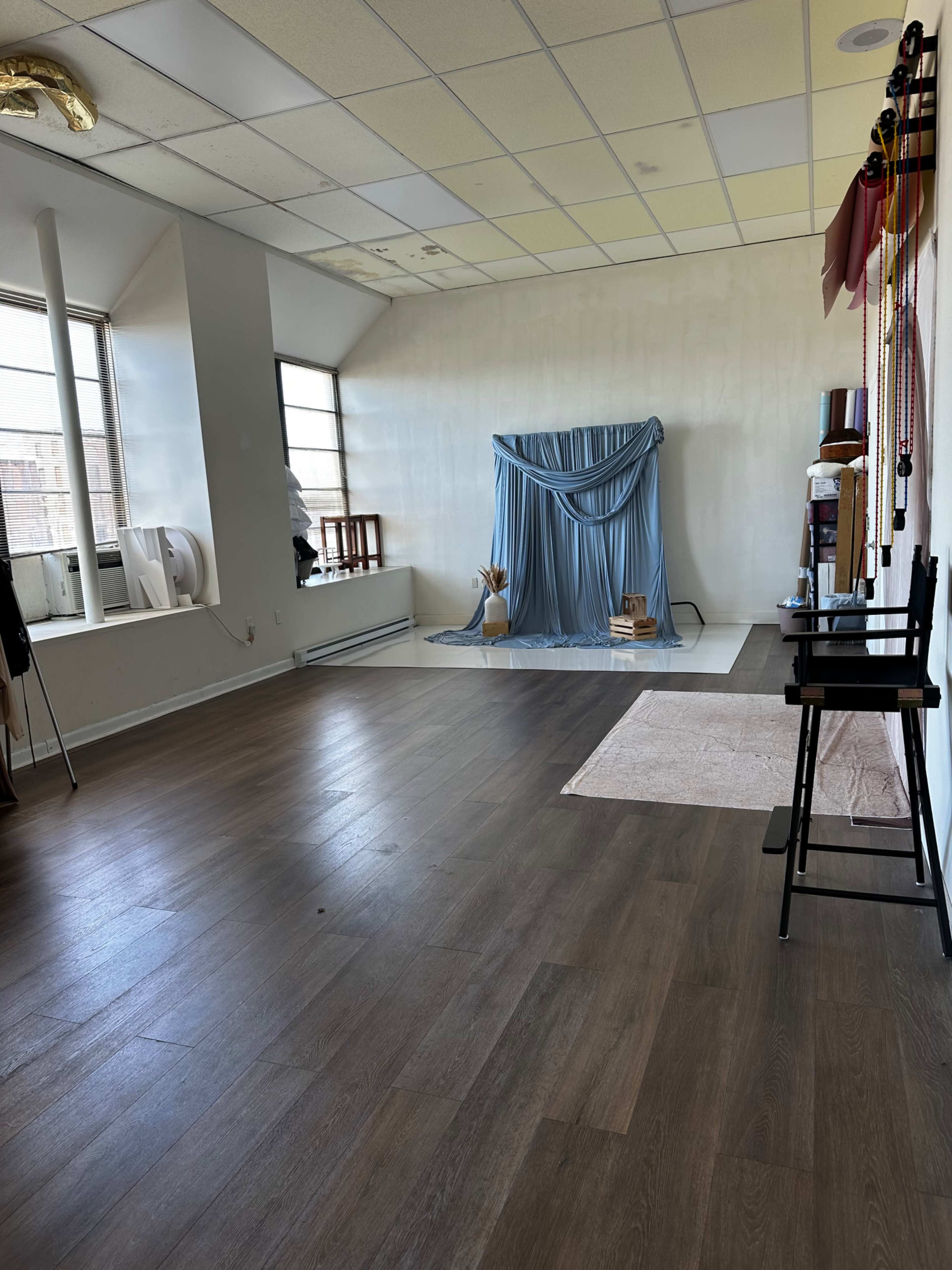 The image shows an empty room with wooden flooring, large windows, and a backdrop draped in blue fabric set up against one wall.