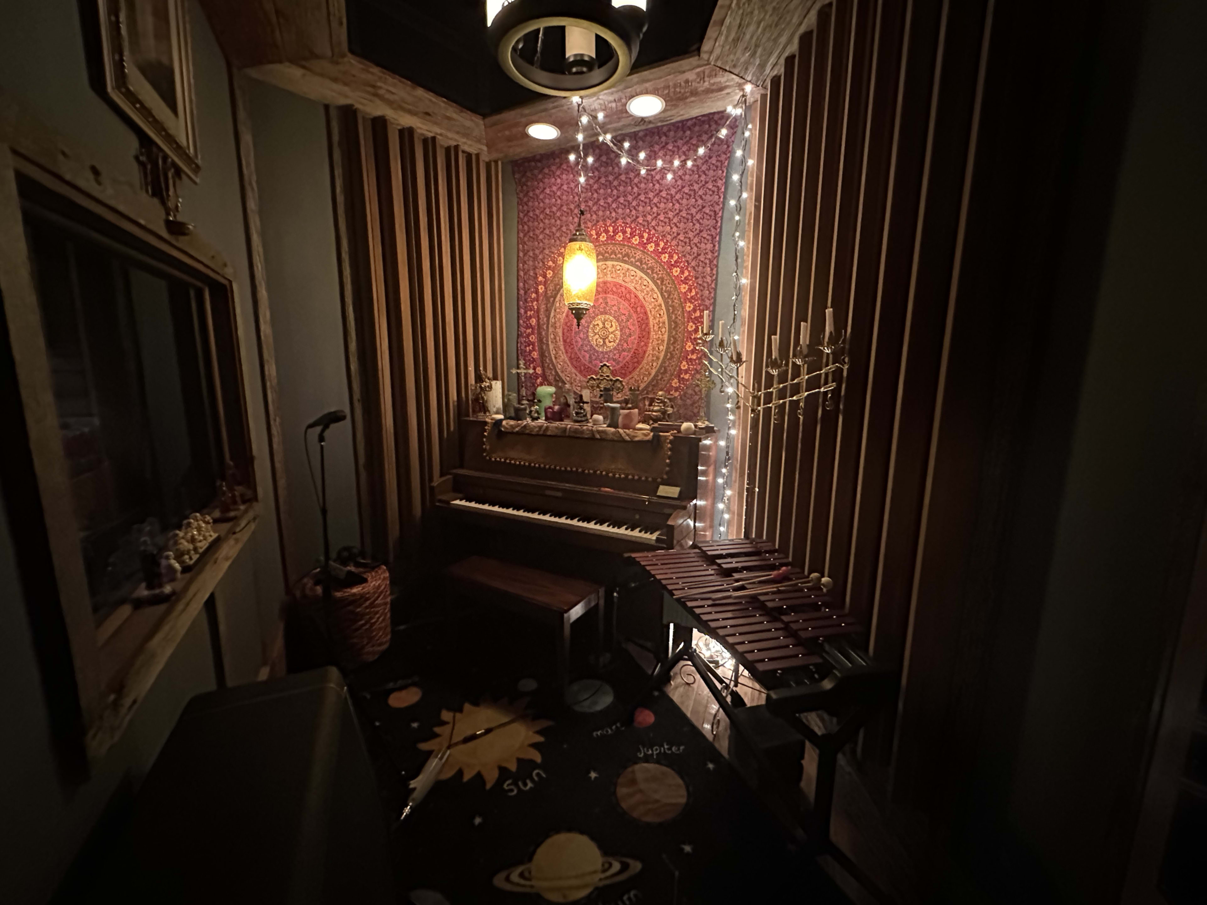 The image shows a cozy music room featuring a piano, a xylophone, and decorative elements, with a tapestry and string lights on the walls.