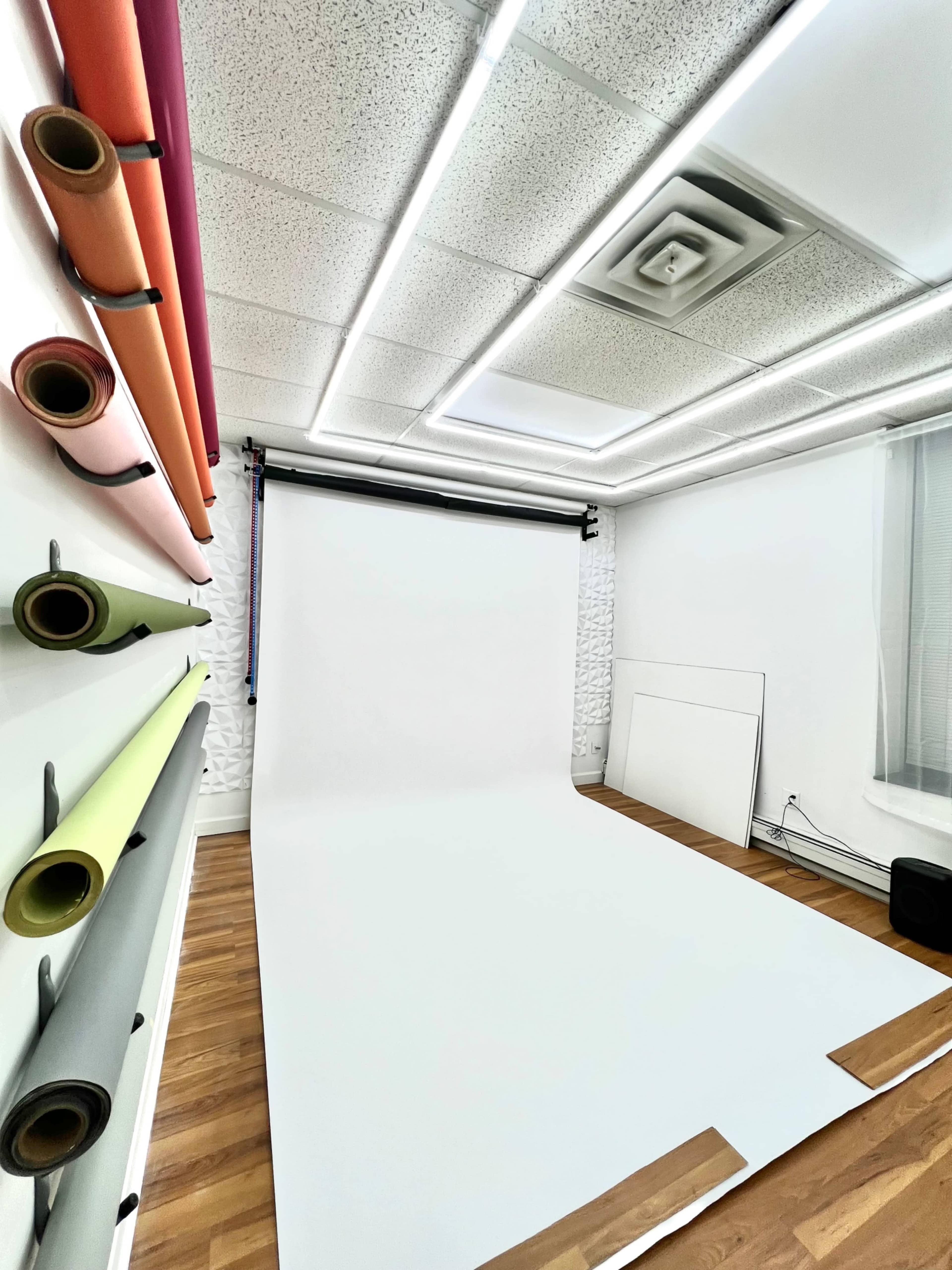 The image shows a photo studio with a white backdrop, wooden flooring, and rolls of colorful backdrop paper mounted on the wall.