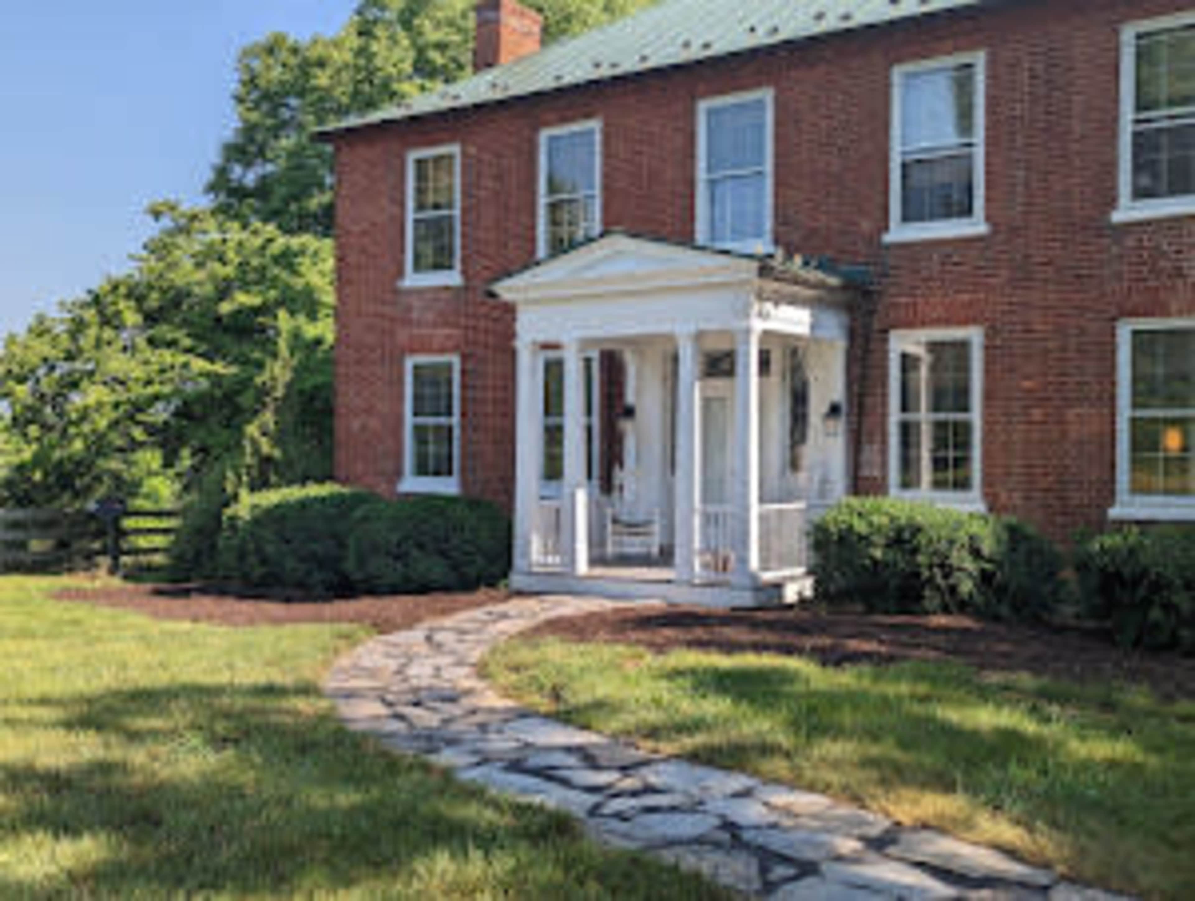 A brick house with a covered front porch is surrounded by well-manicured grass and shrubs, connected by a stone pathway.