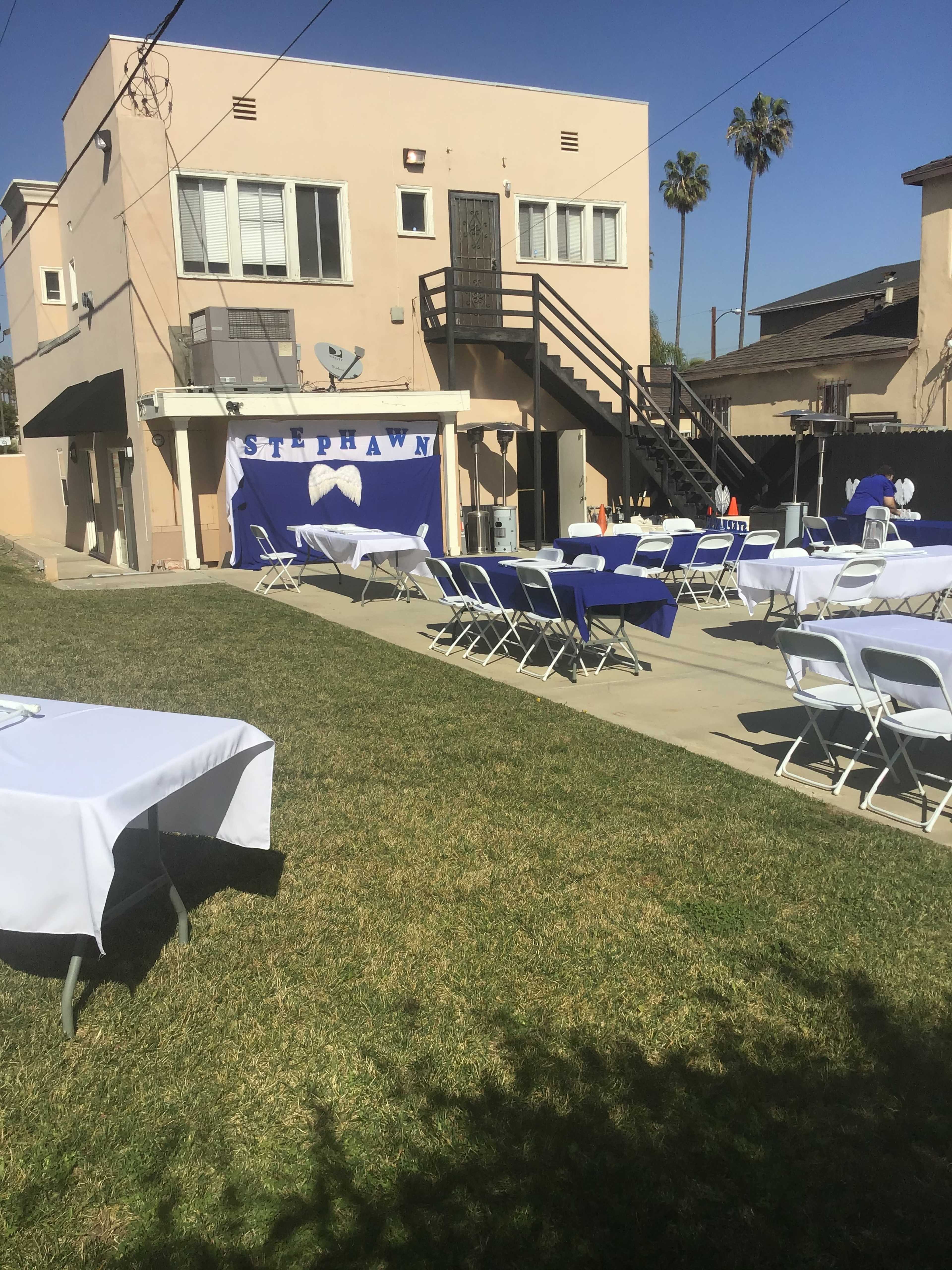 A backyard setup for an event with several white tables covered with blue tablecloths and a backdrop featuring the name "STEPHAWN" on a wall.