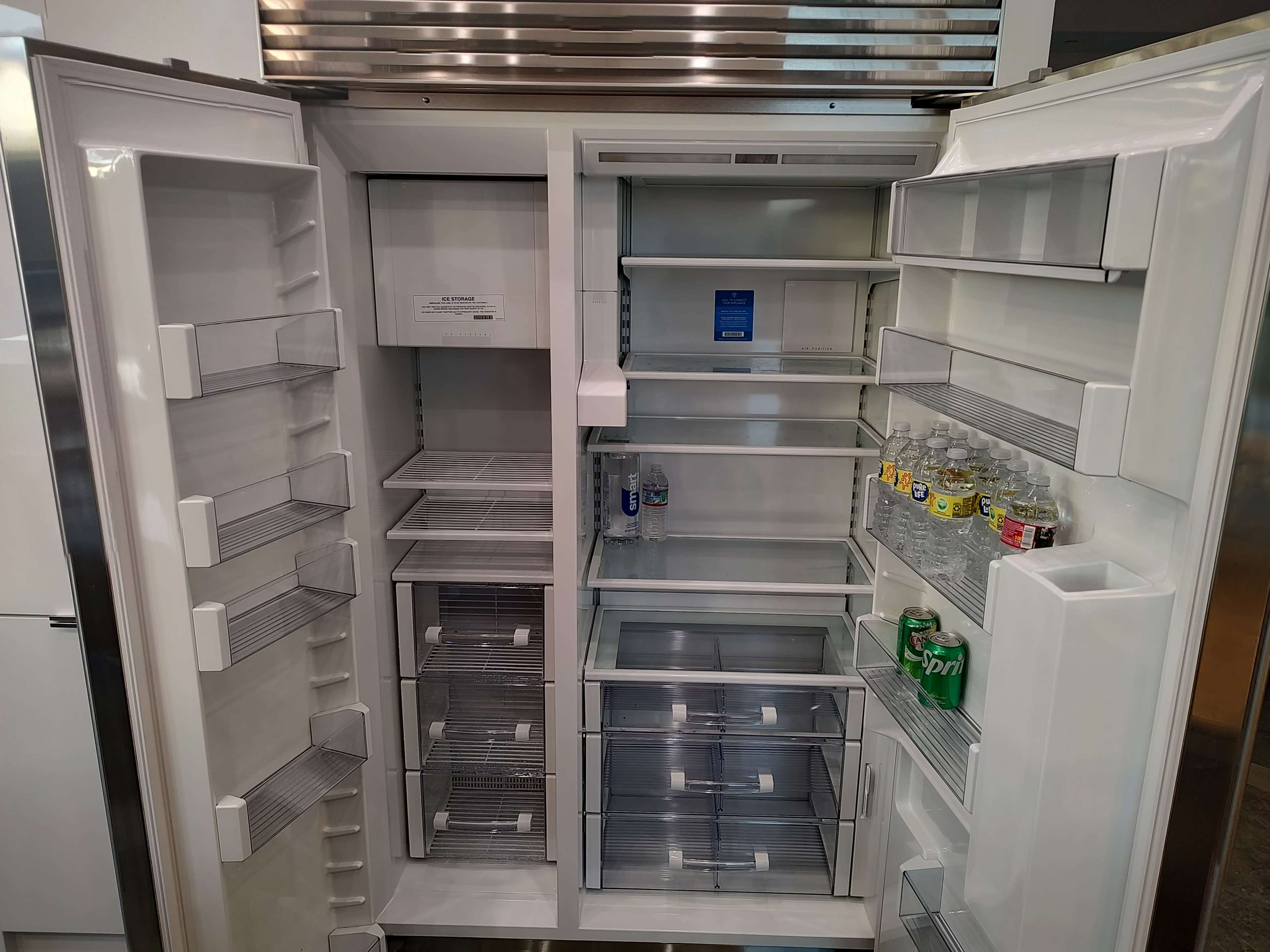 An open refrigerator with several shelves, drawers, and compartments that are mostly empty, displaying a few bottles and cans.