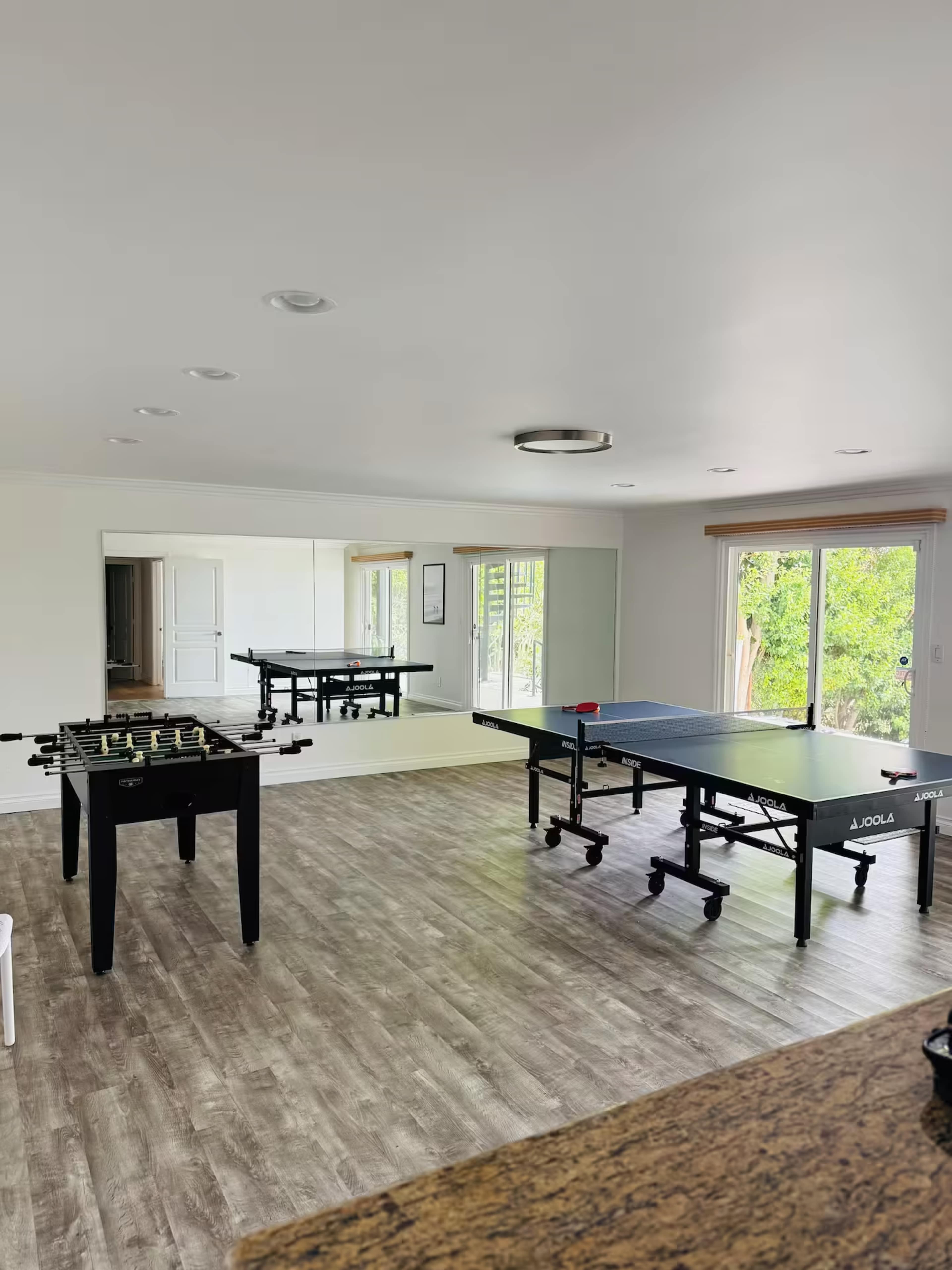 The image shows a bright, spacious room featuring a foosball table and two ping pong tables, with large windows providing natural light.