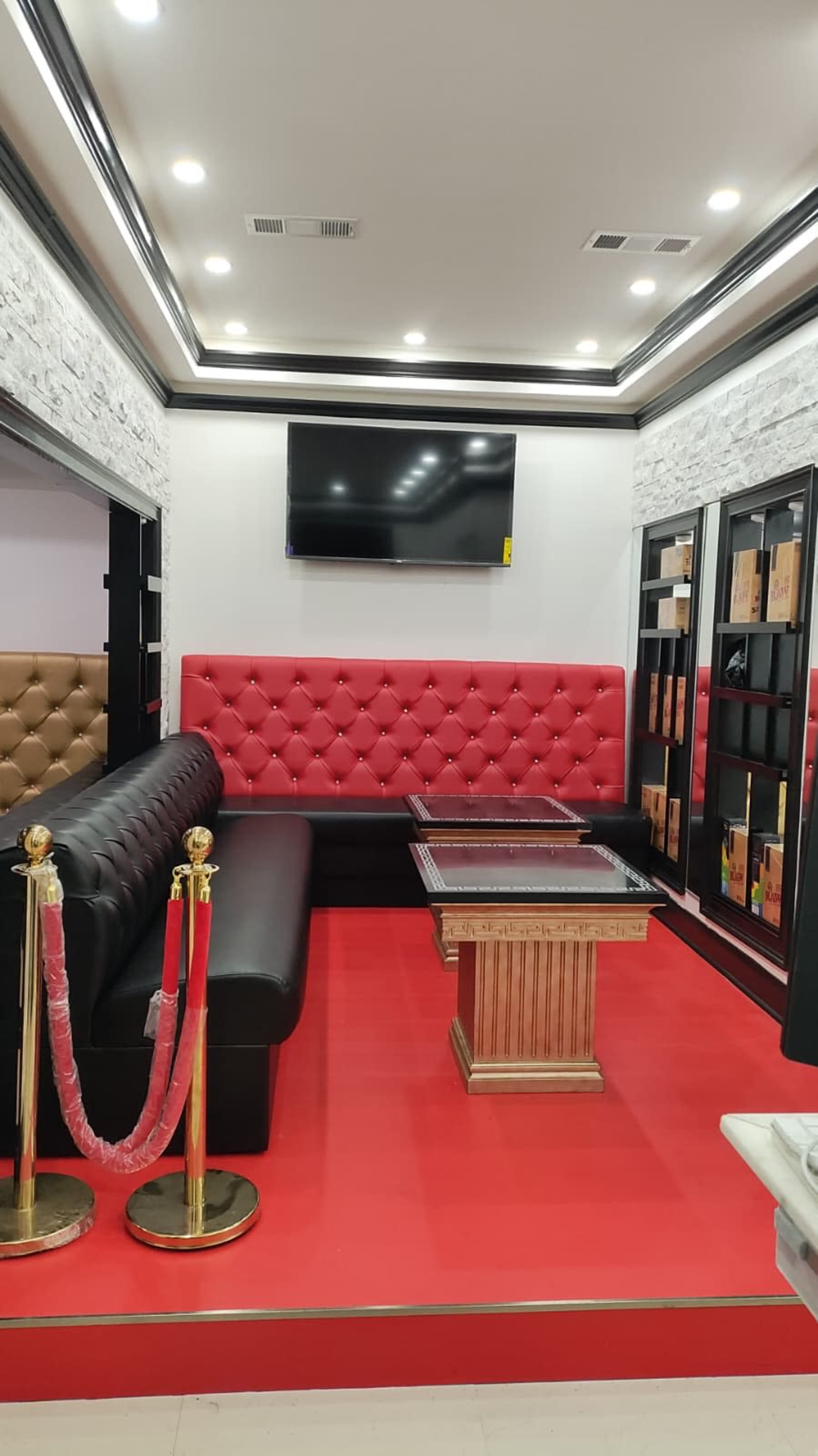 The image shows a modern interior of a lounge area featuring a red tufted bench, black leather seating, a glass-topped table, and a large TV mounted on the wall.