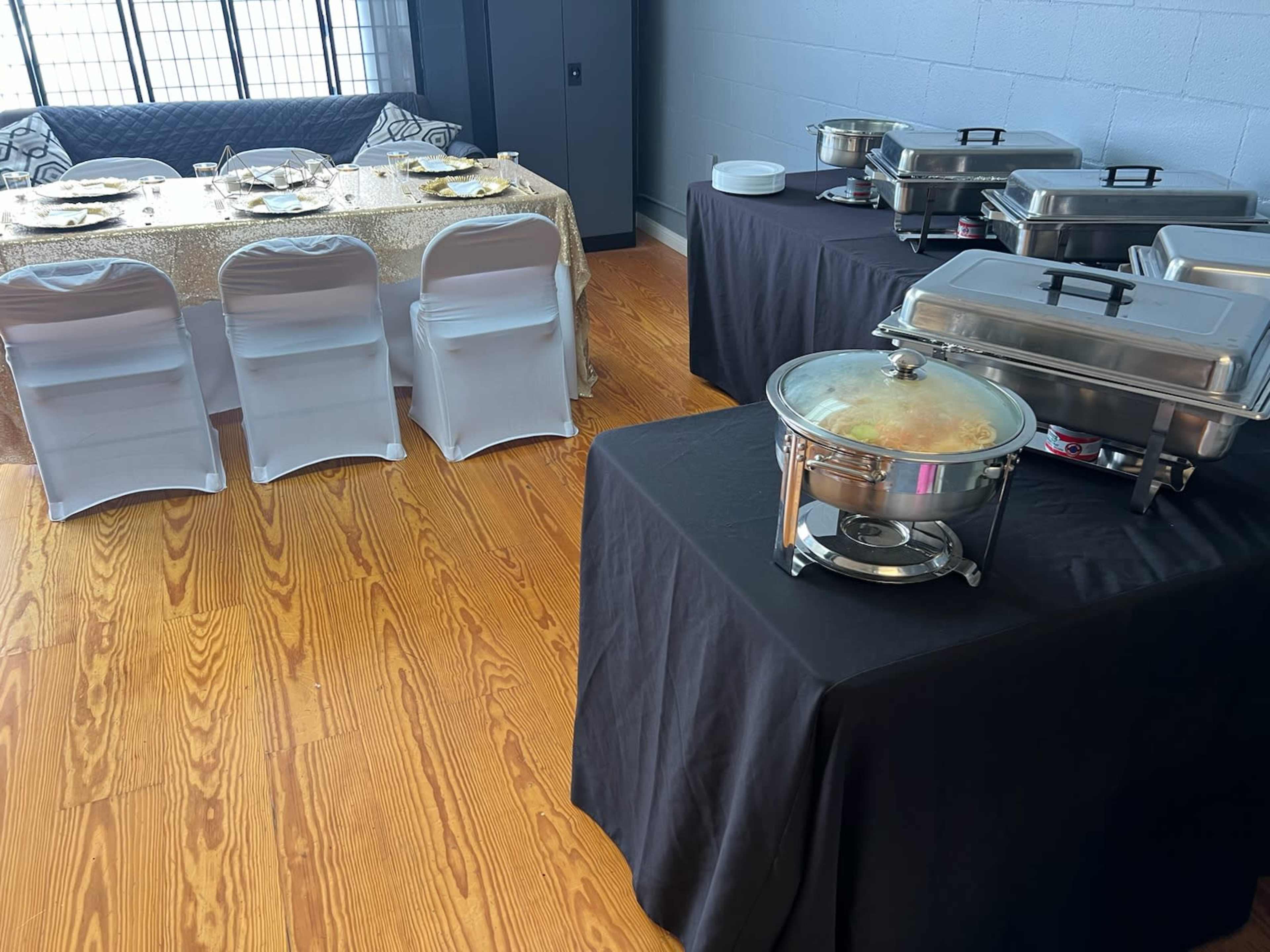 The image shows a dining area with a table set for a meal, featuring gold tablecloths and stylish chairs, alongside black tables with food warmers.