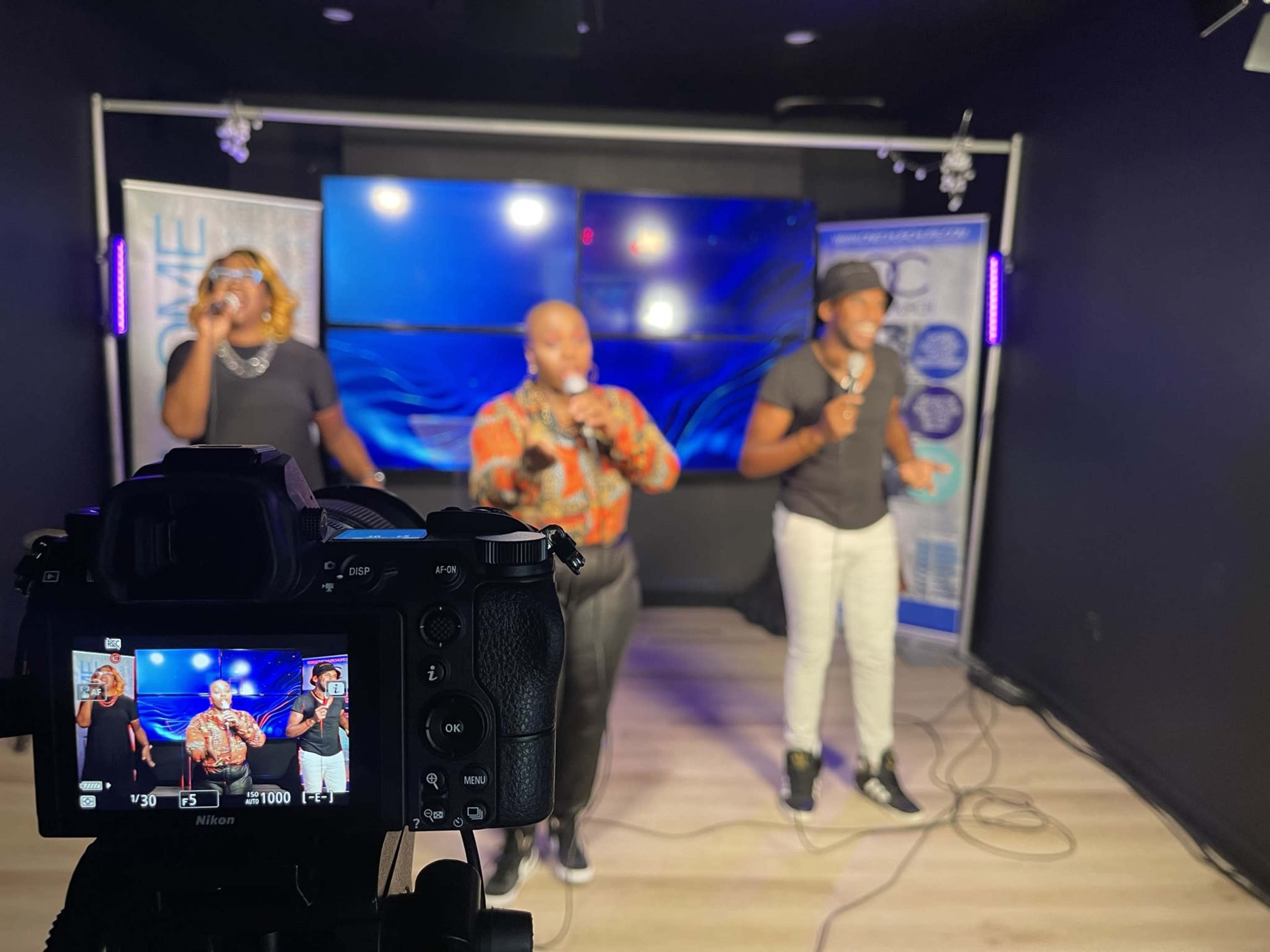Three performers sing into microphones on stage while a camera captures the scene.