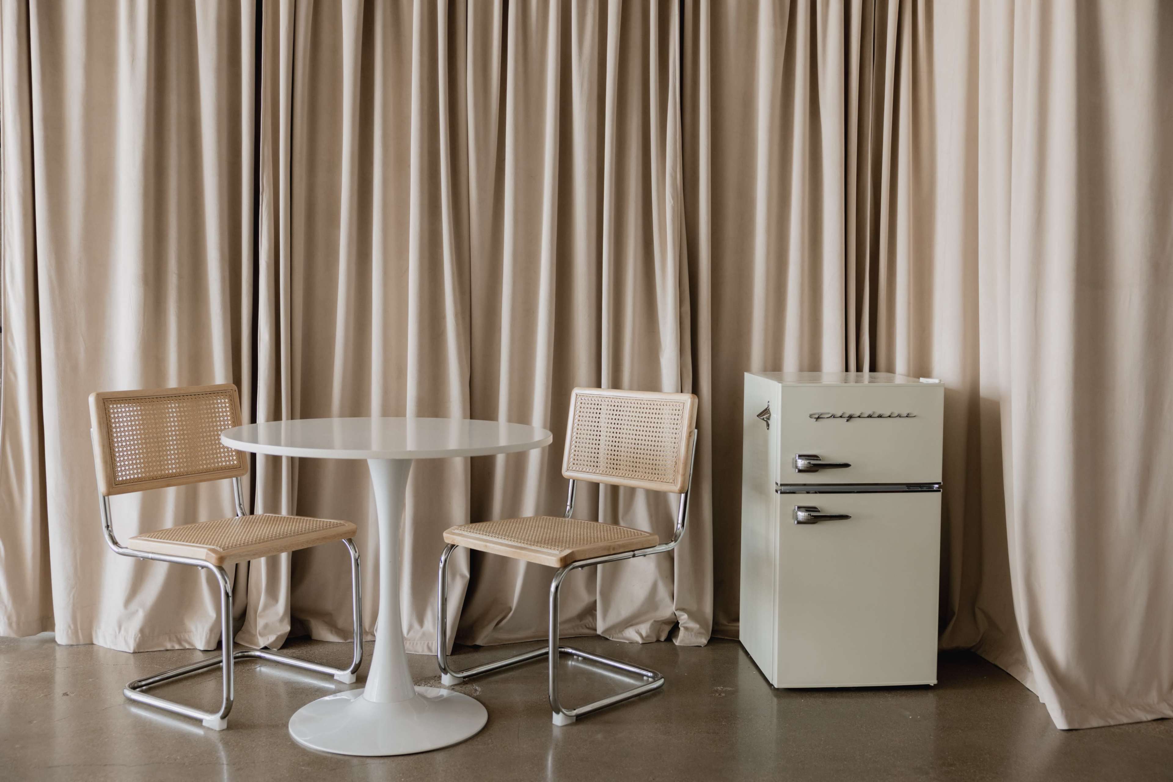 A round white table and two woven chairs are positioned next to a vintage-style cream refrigerator against a backdrop of beige curtains.