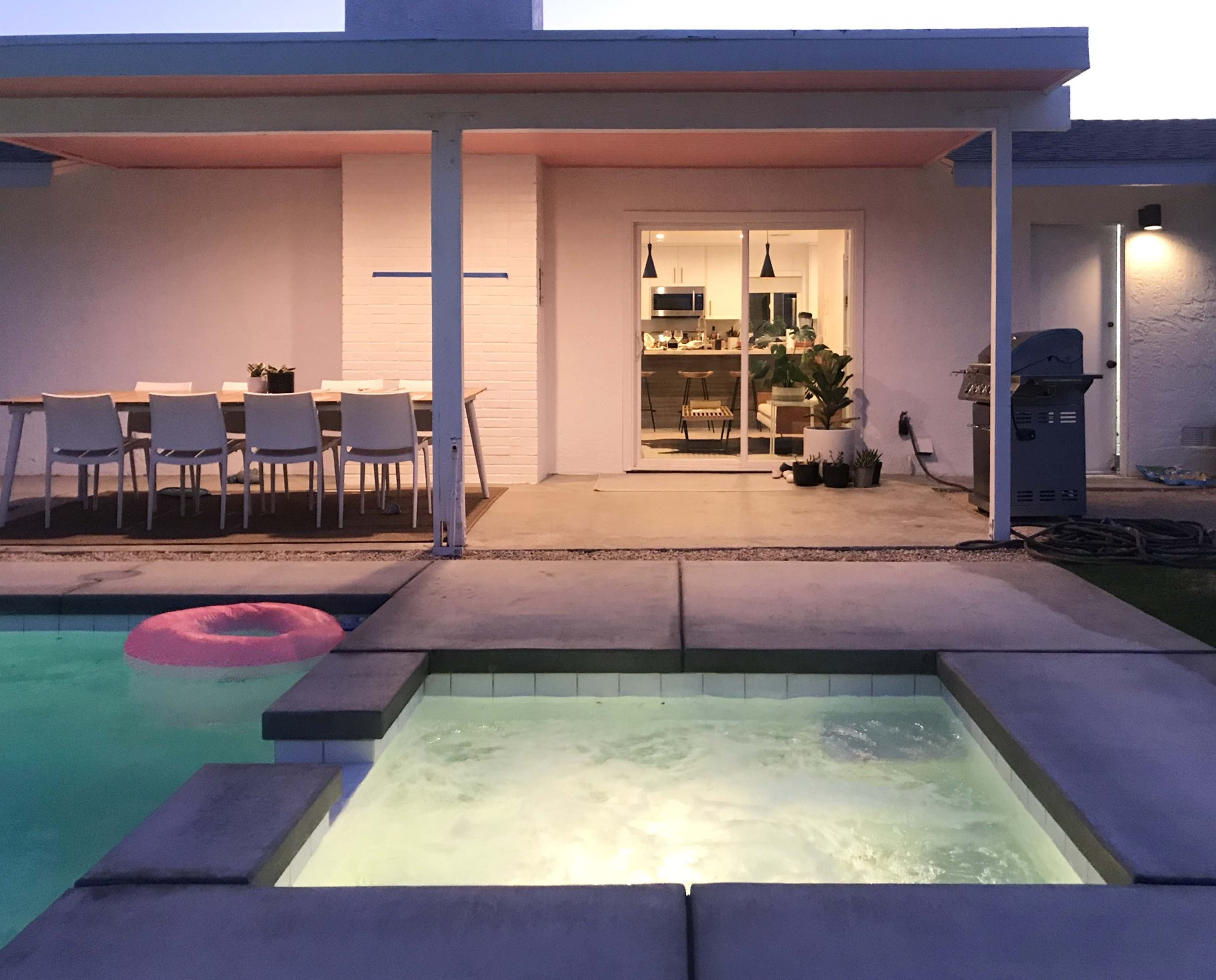 The image shows a modern outdoor space featuring a swimming pool, a hot tub, and a dining area with white chairs and a table, set against a minimalist house facade at dusk.