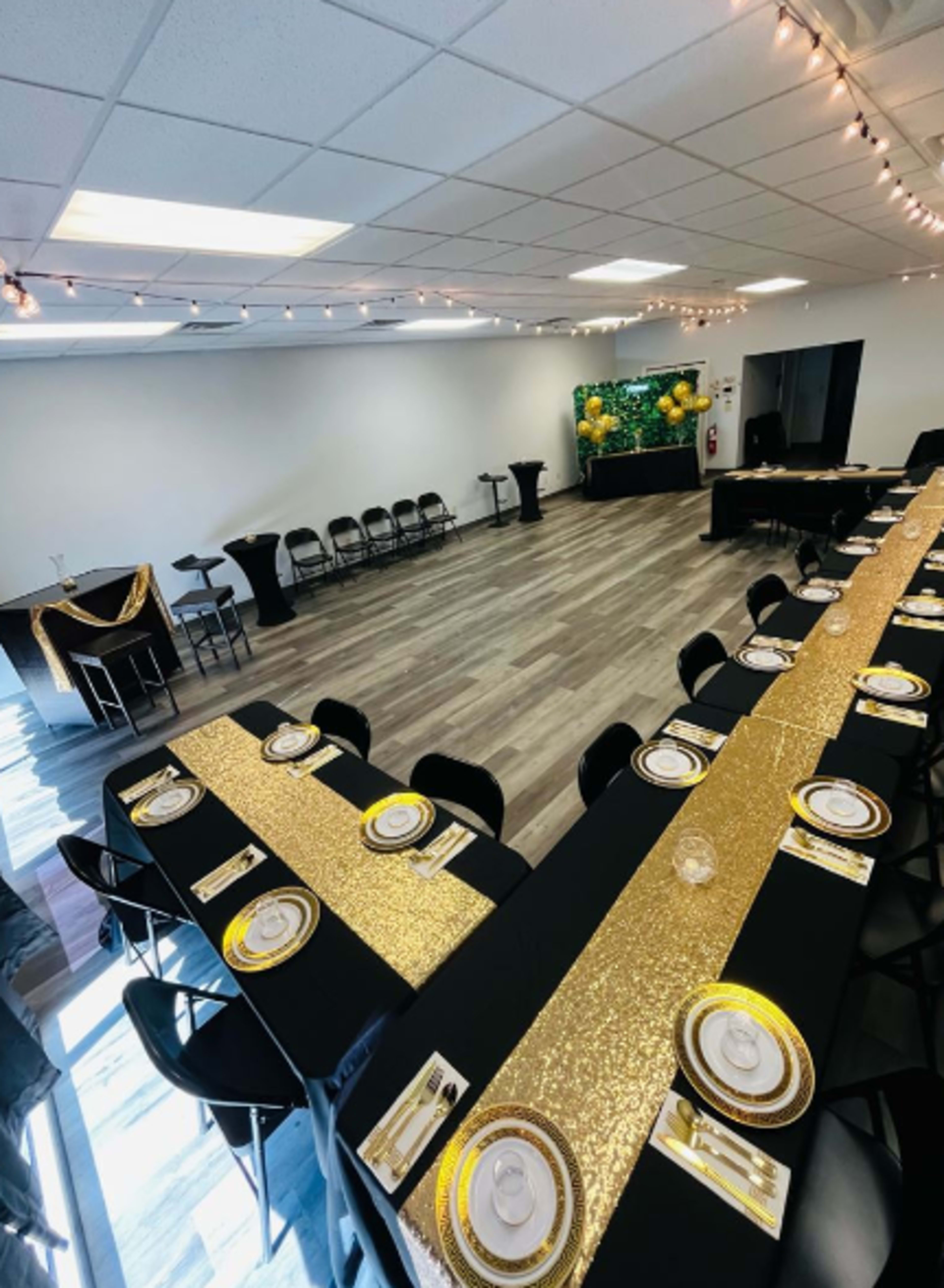 A large event space is set up with a long black table adorned with a gold sequined runner and plates, surrounded by chairs, and decorated with string lights.