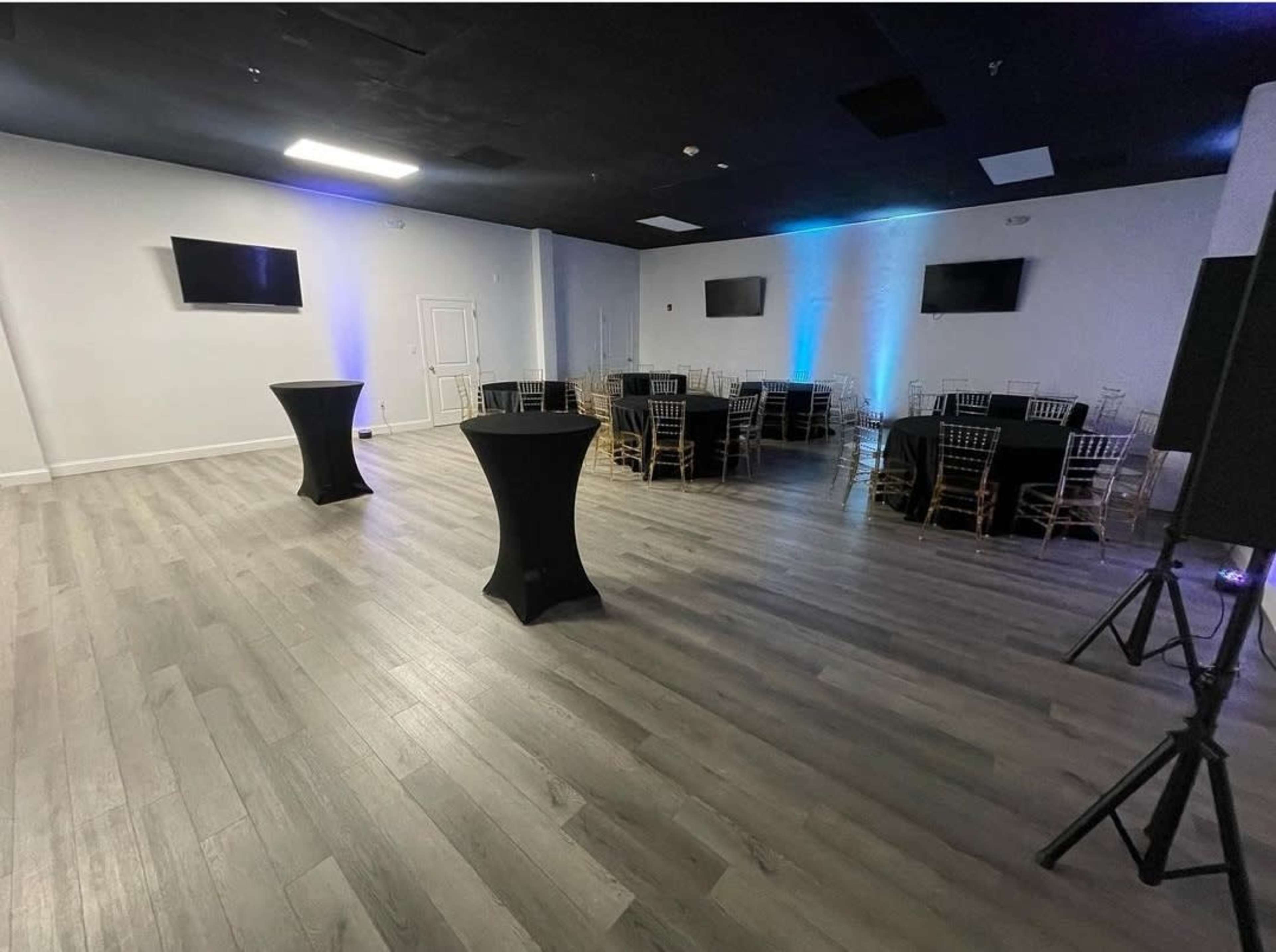 A spacious event room features black tables, gold chairs, and two wall-mounted TVs.