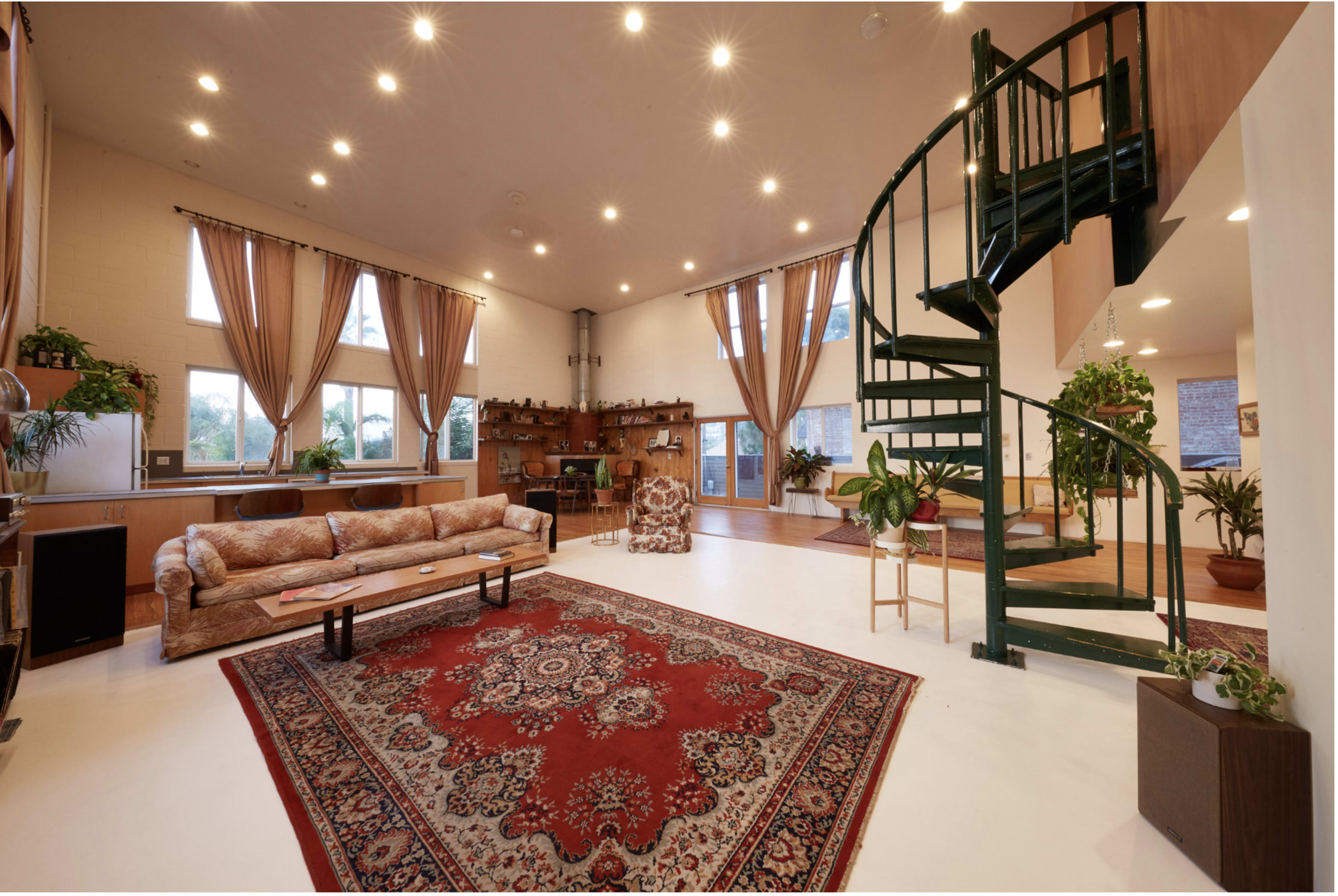 A spacious living room with large windows, a patterned rug, a spiral staircase, and several potted plants.