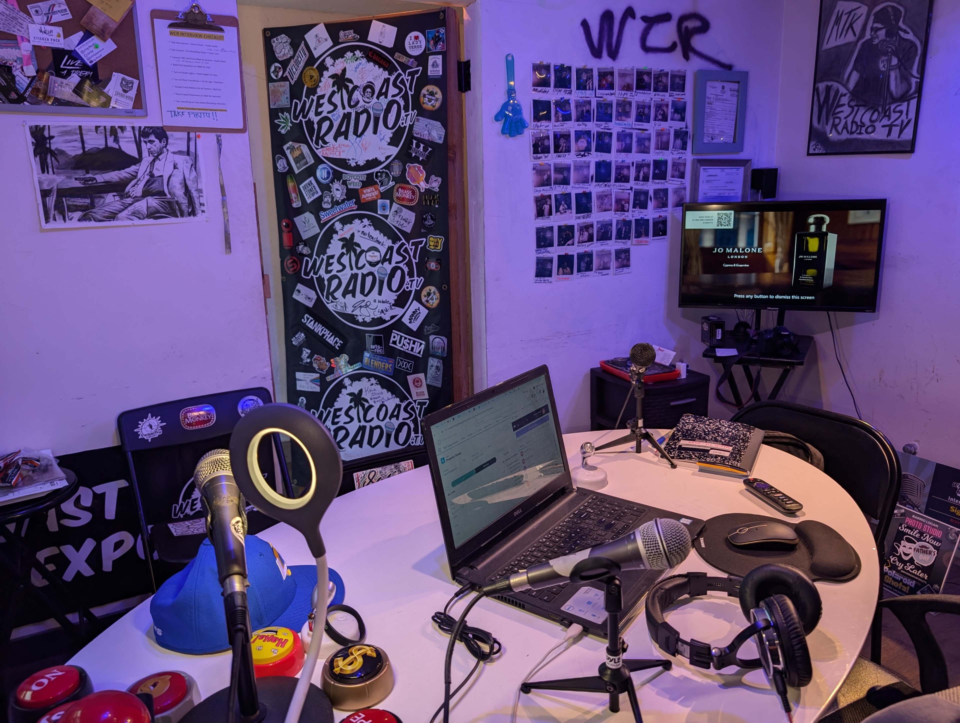 A small, cluttered radio studio with microphones, a laptop, a television displaying content, and various stickers on the walls.