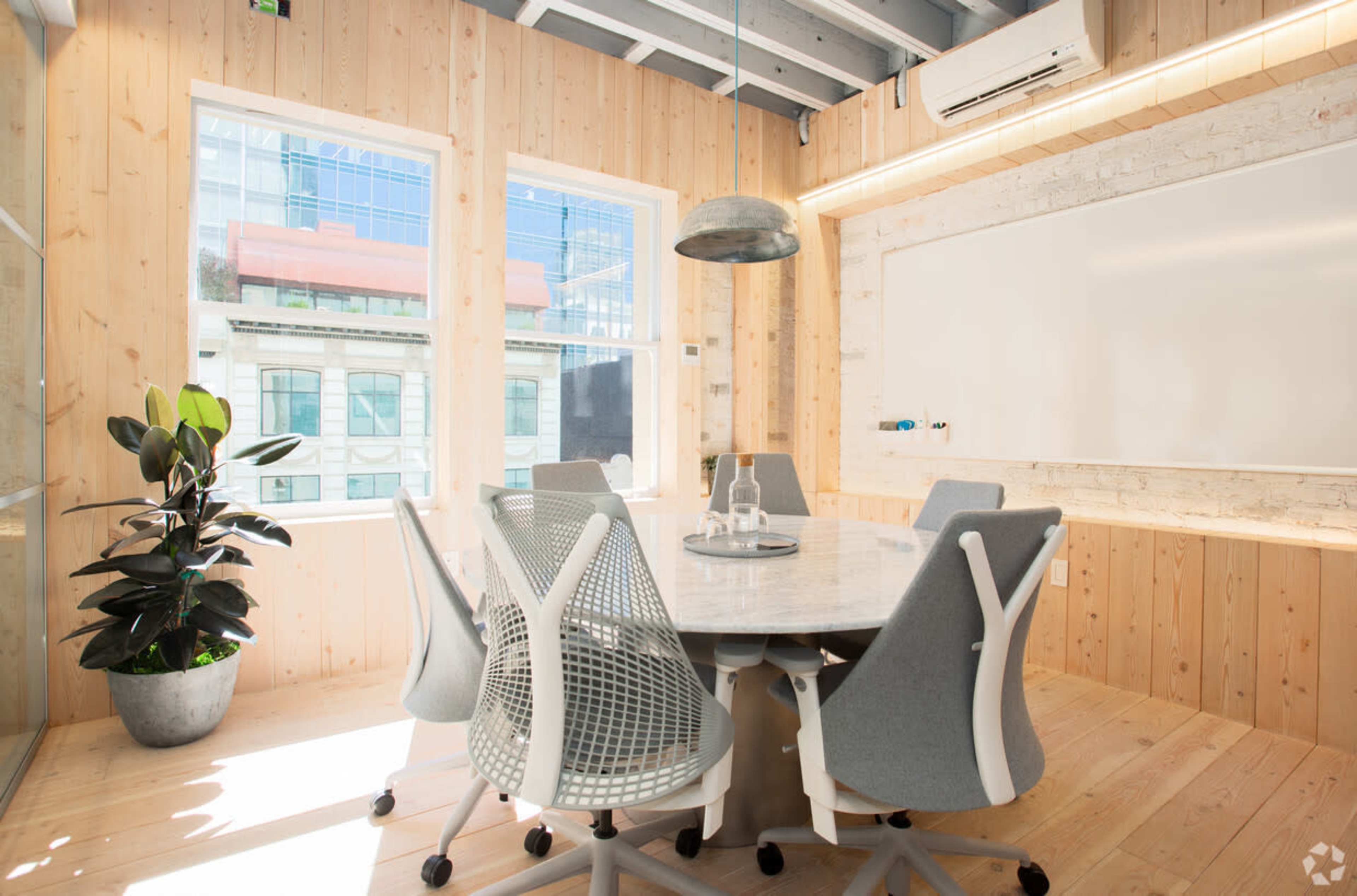 A brightly lit conference room features a round marble table surrounded by six gray office chairs, with large windows that provide natural light and a potted plant in one corner.