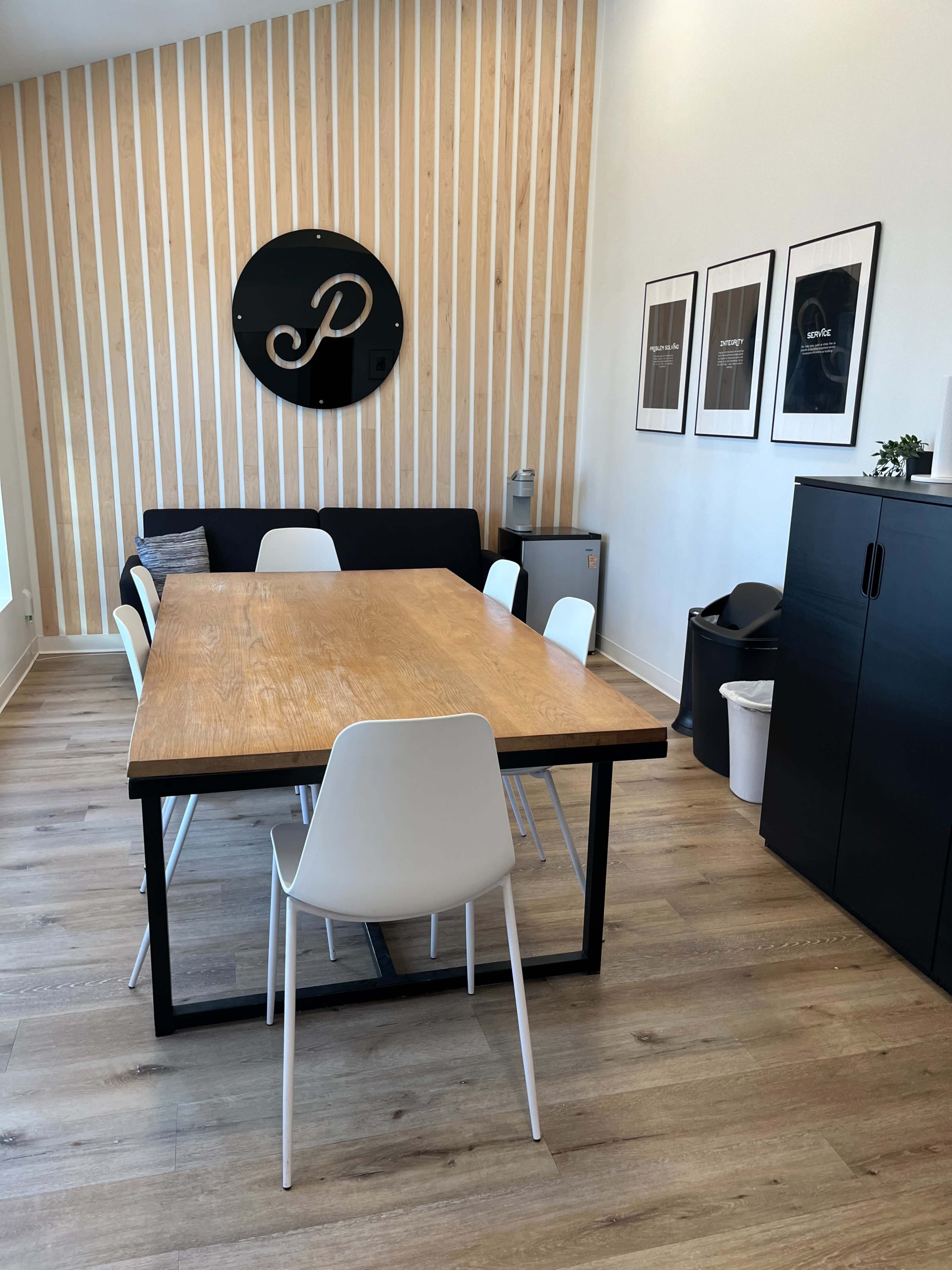 A minimalist dining area features a wooden table surrounded by white chairs against a light wood paneled wall, with framed artwork and a black cabinet nearby.