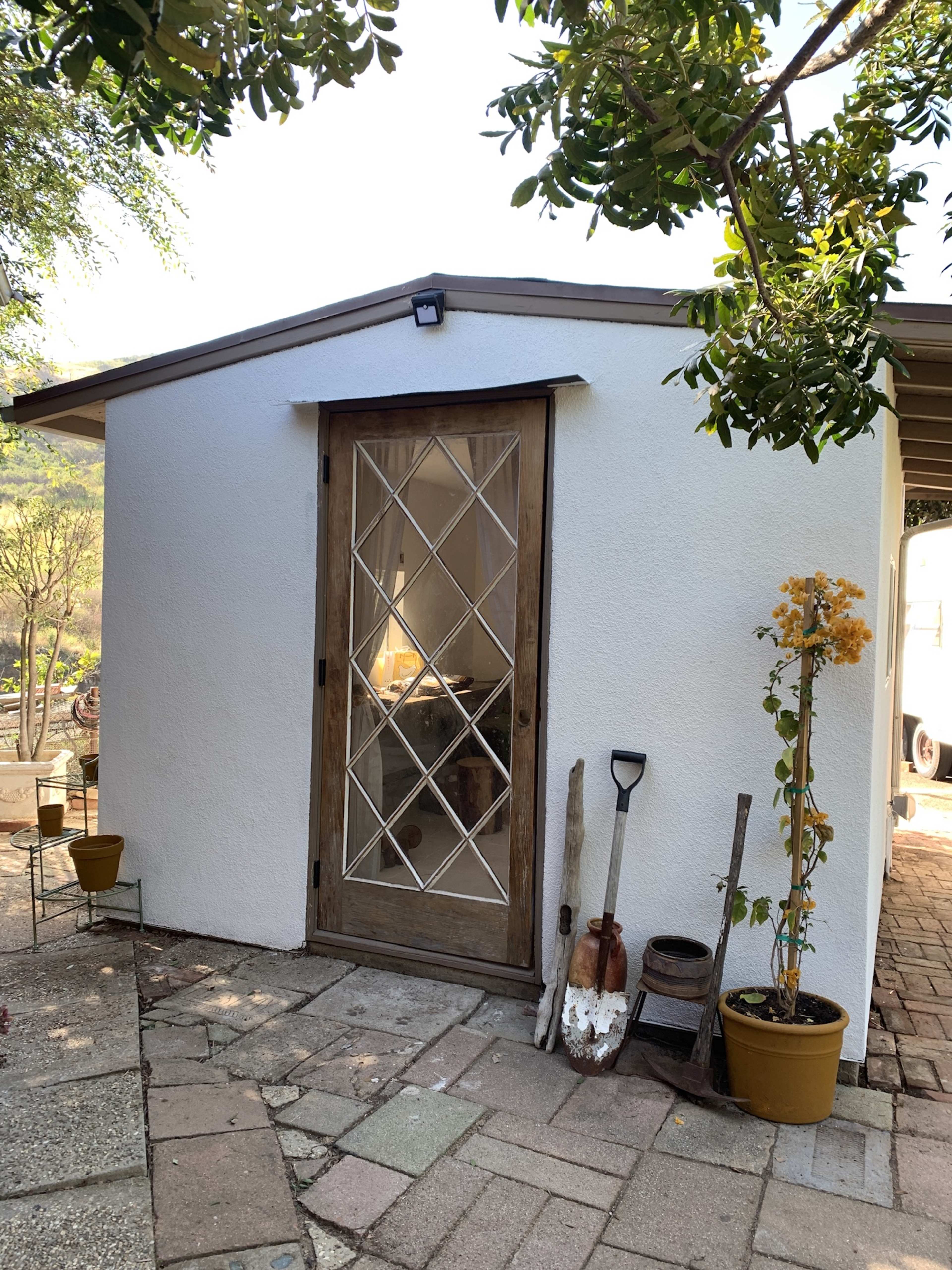 A small white-walled building with a wooden door featuring diamond-shaped glass panes, surrounded by potted plants and garden tools on a stone pathway.