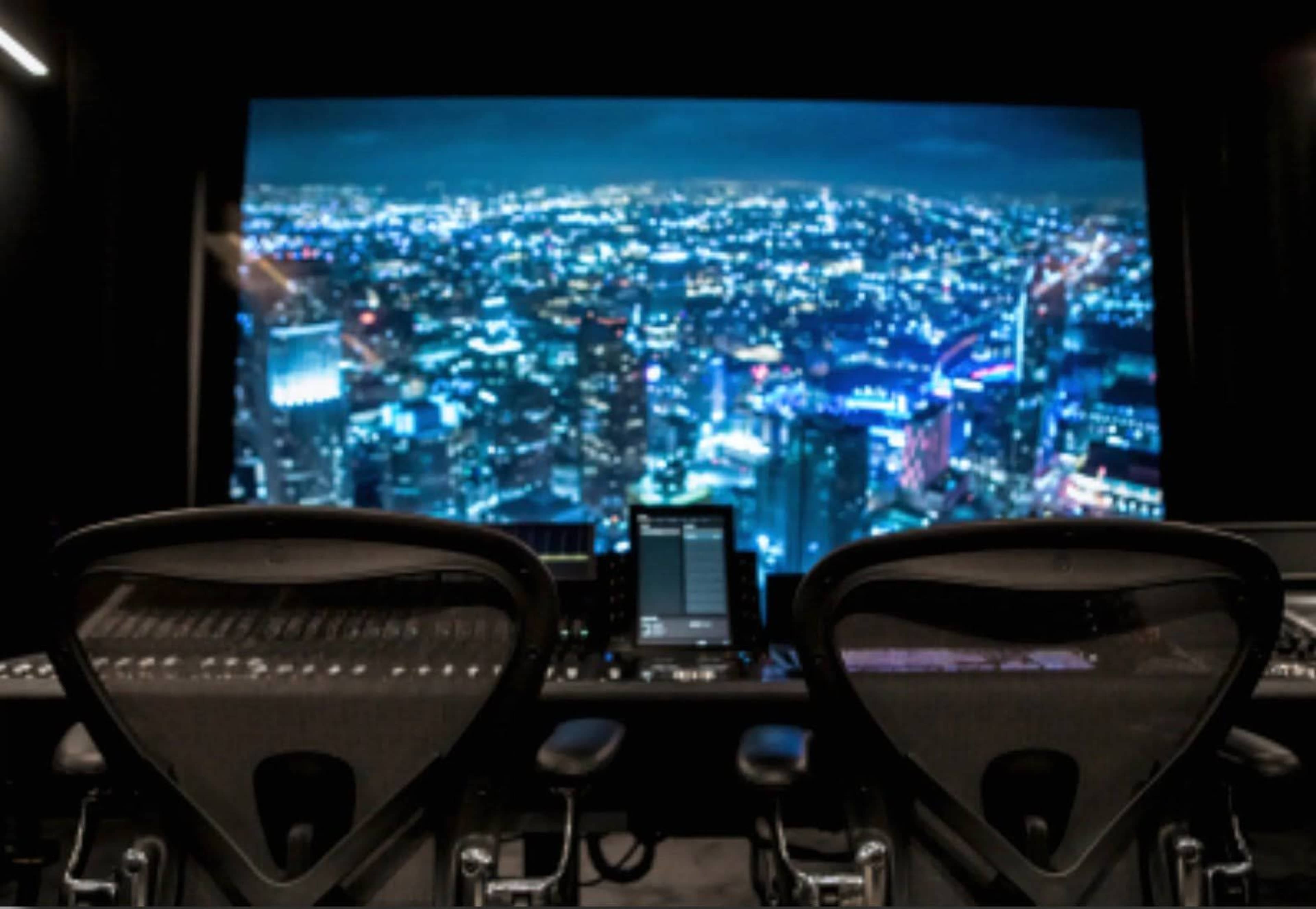 The image shows two black chairs facing a large screen displaying a panoramic view of a city skyline at night, illuminated by numerous lights.