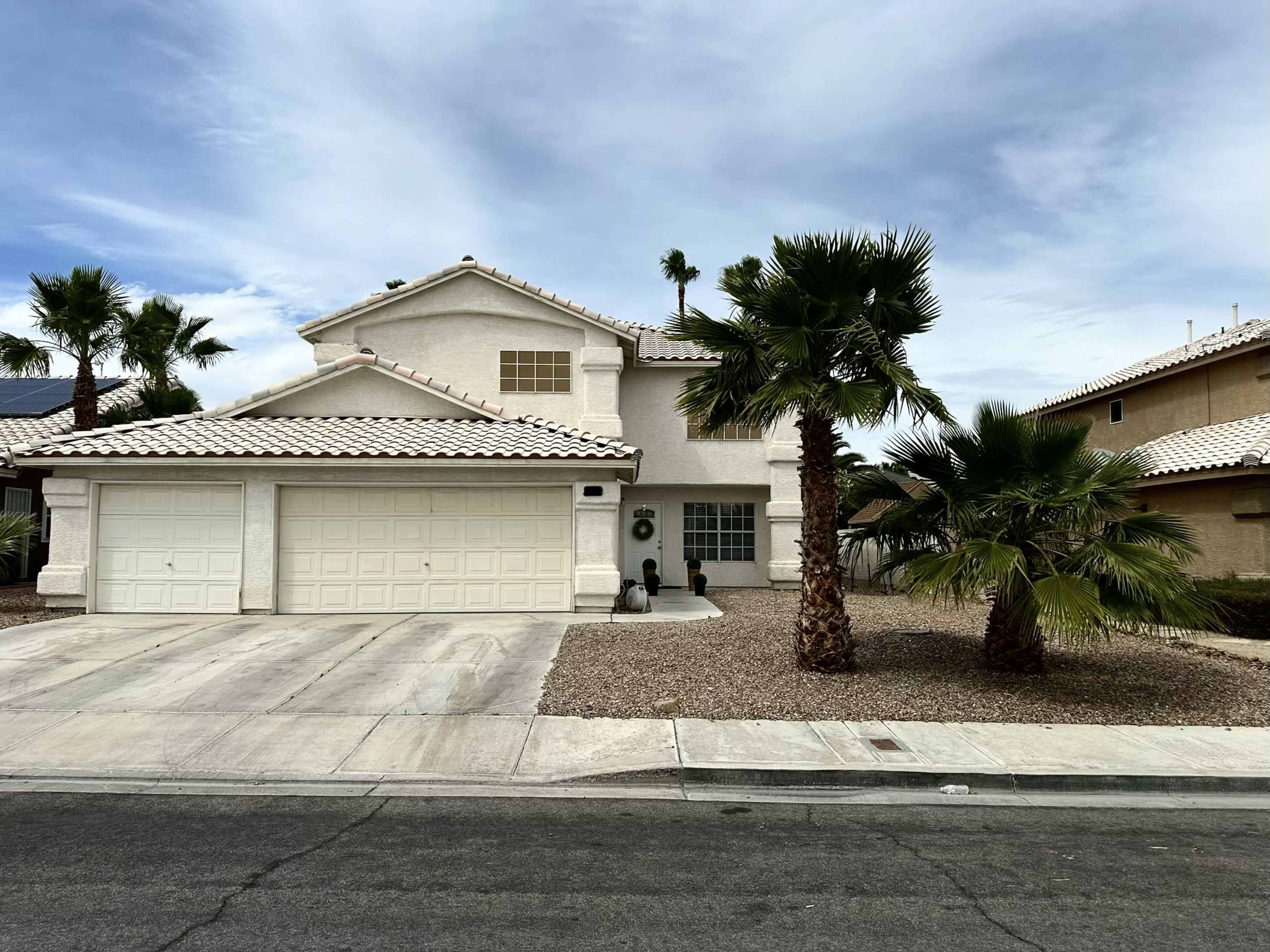 The image shows a two-story house with a tile roof and a two-car garage, surrounded by palm trees and gravel landscaping.