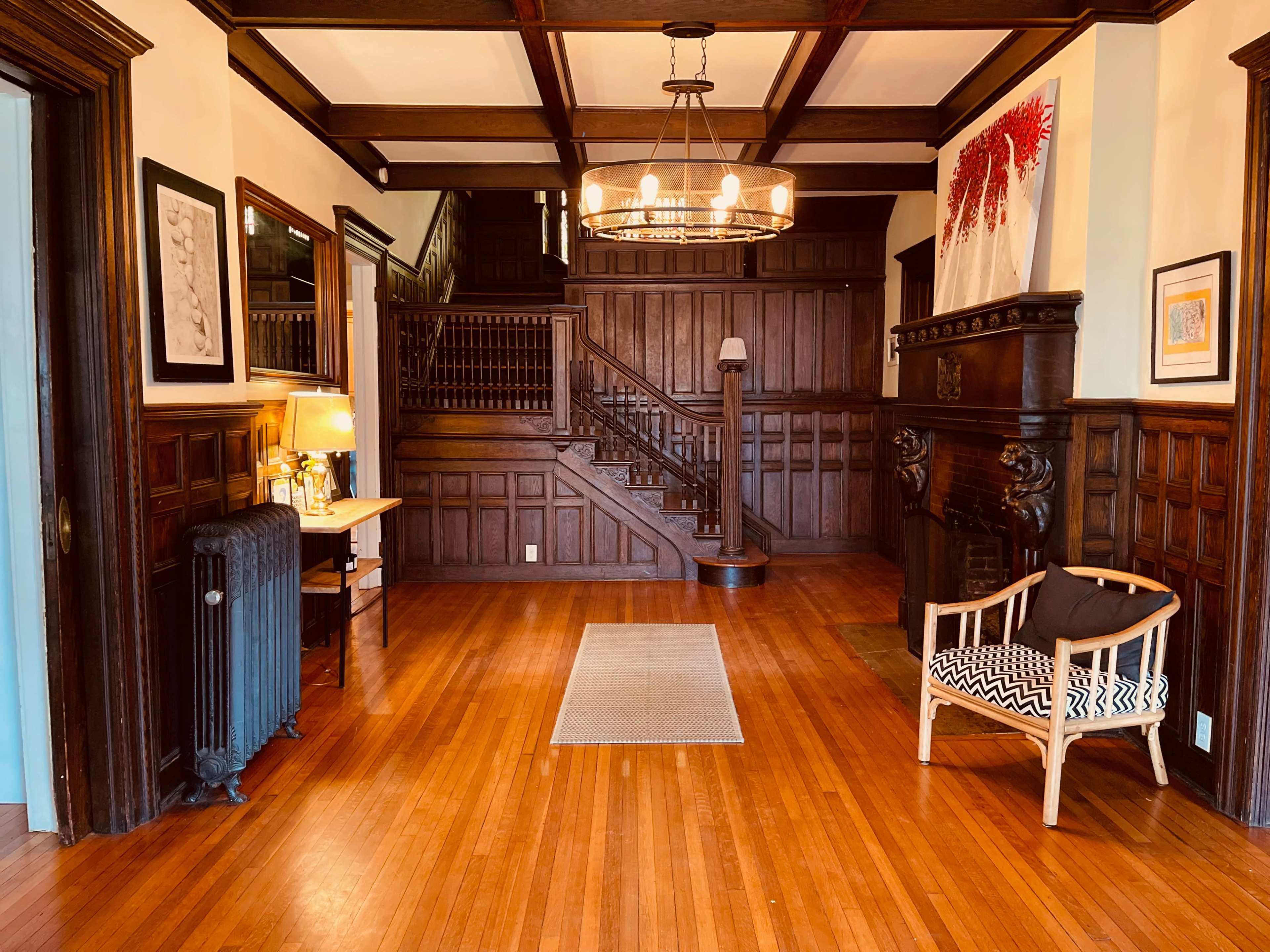 A large hallway featuring wooden paneling, a staircase with a decorative railing, a chandelier, and a cozy seating area.