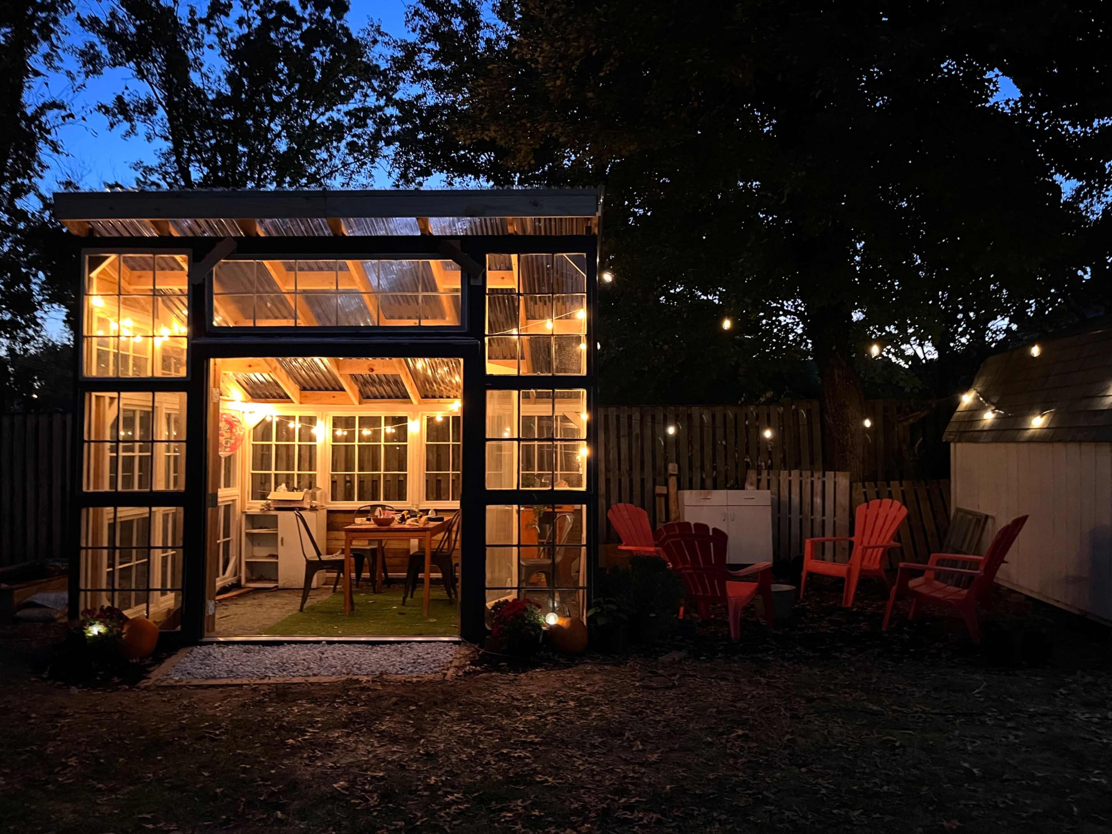 A glass greenhouse with warm lights is illuminated at dusk, surrounded by red outdoor chairs and a wooden fence.