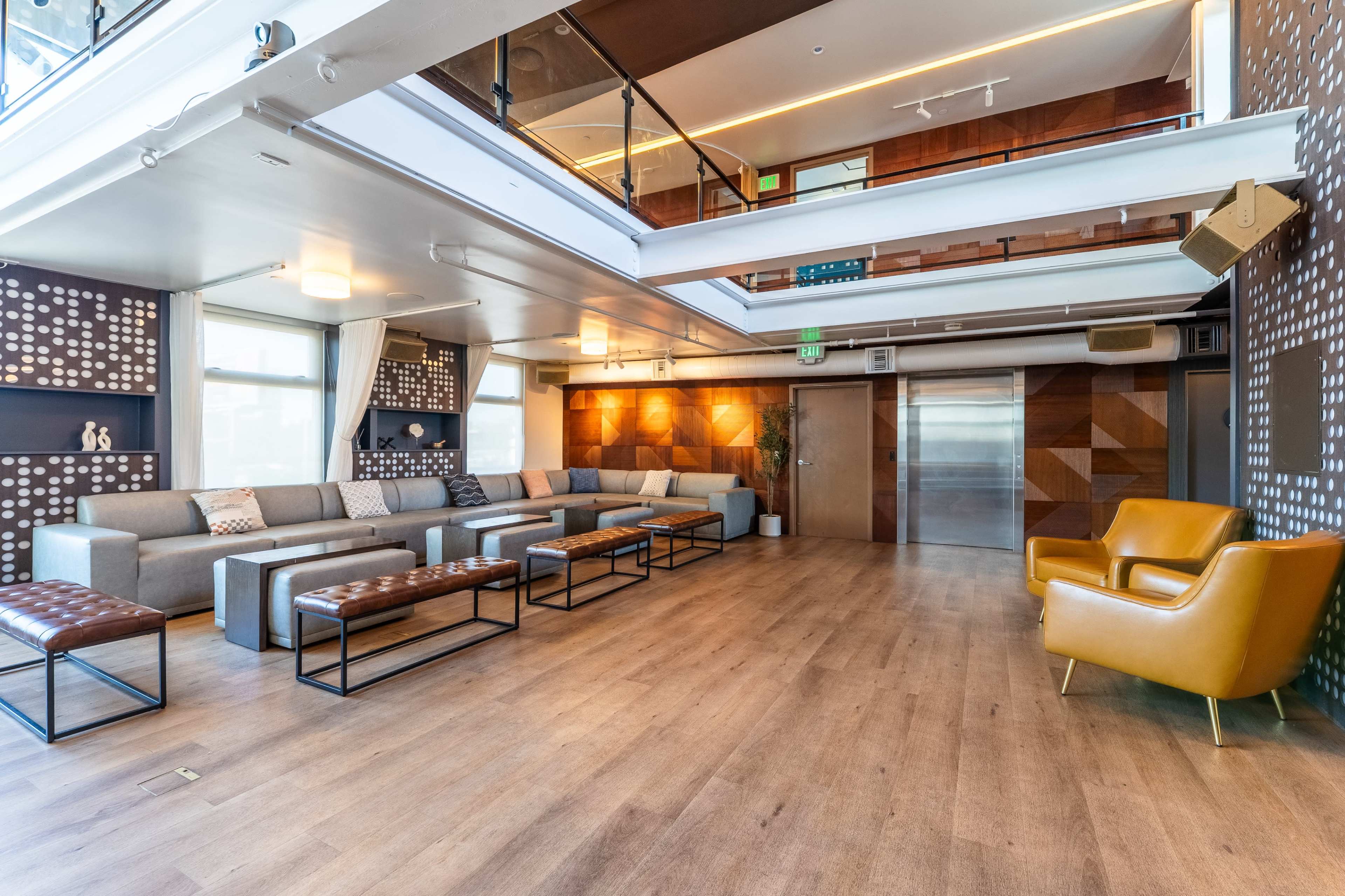 The image shows a modern lounge area with gray sofas, leather ottomans, and a yellow armchair, featuring wooden flooring and large windows.