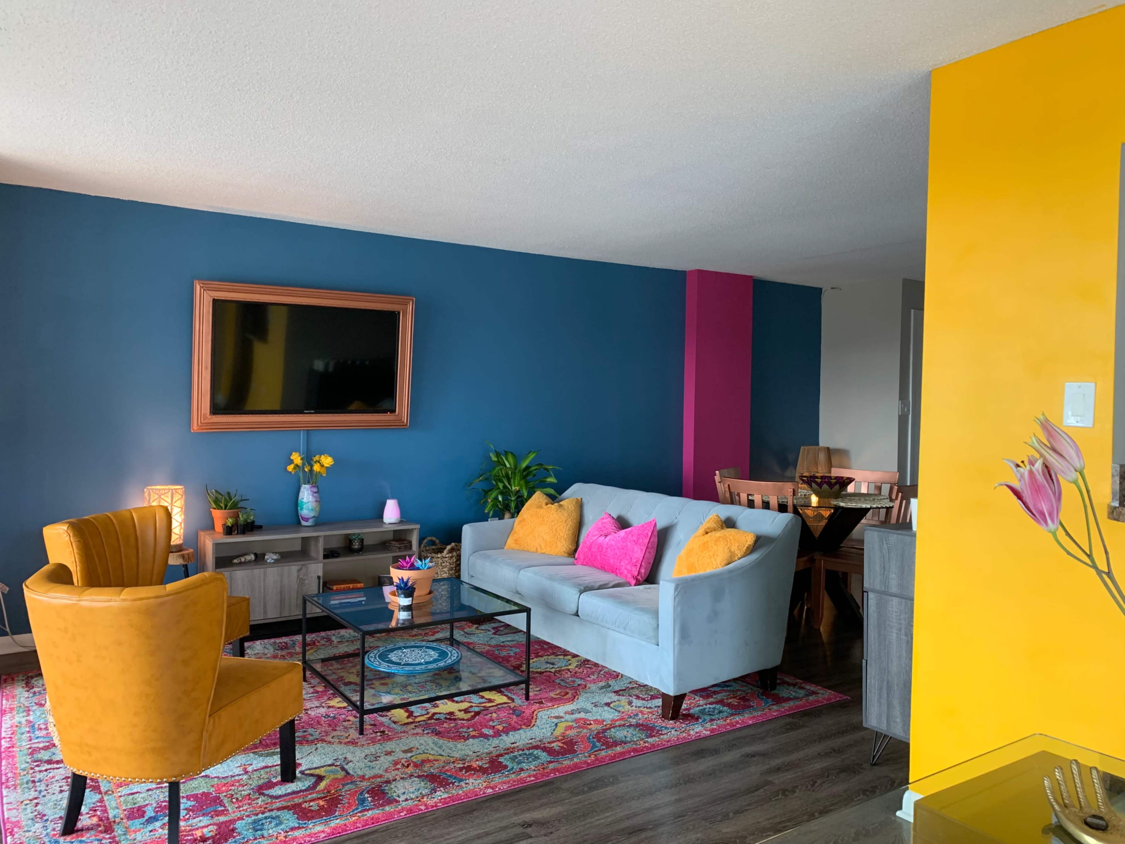 The living room features a blue accent wall, a gray couch, yellow chairs, a coffee table, and a patterned area rug, with a dining area visible in the background.