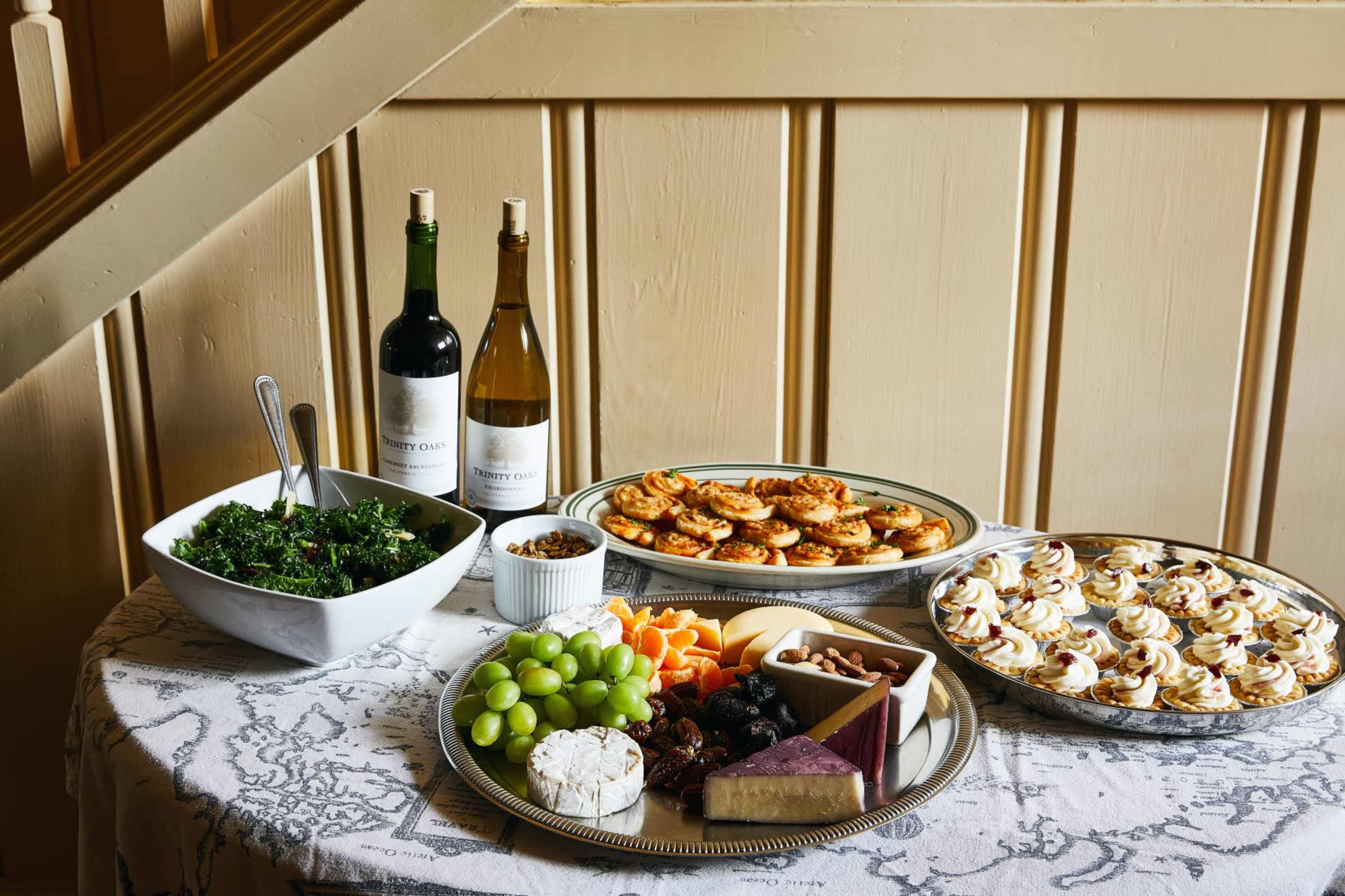A table is set with a variety of foods, including salads, cheeses, fruits, and pastries, alongside bottles of red and white wine.