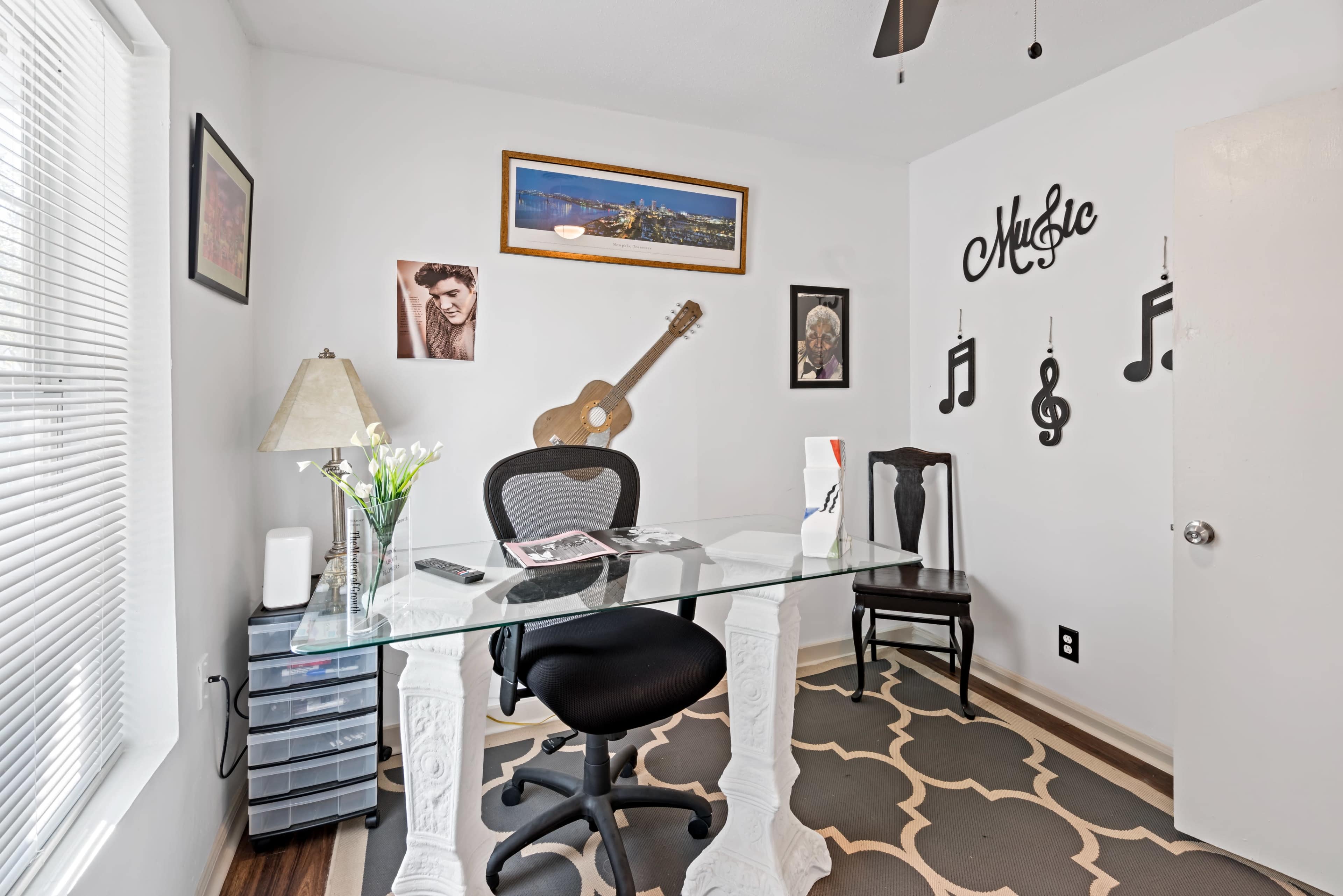 A home office features a glass desk, two chairs, a lamp, and musical decor against a white wall.