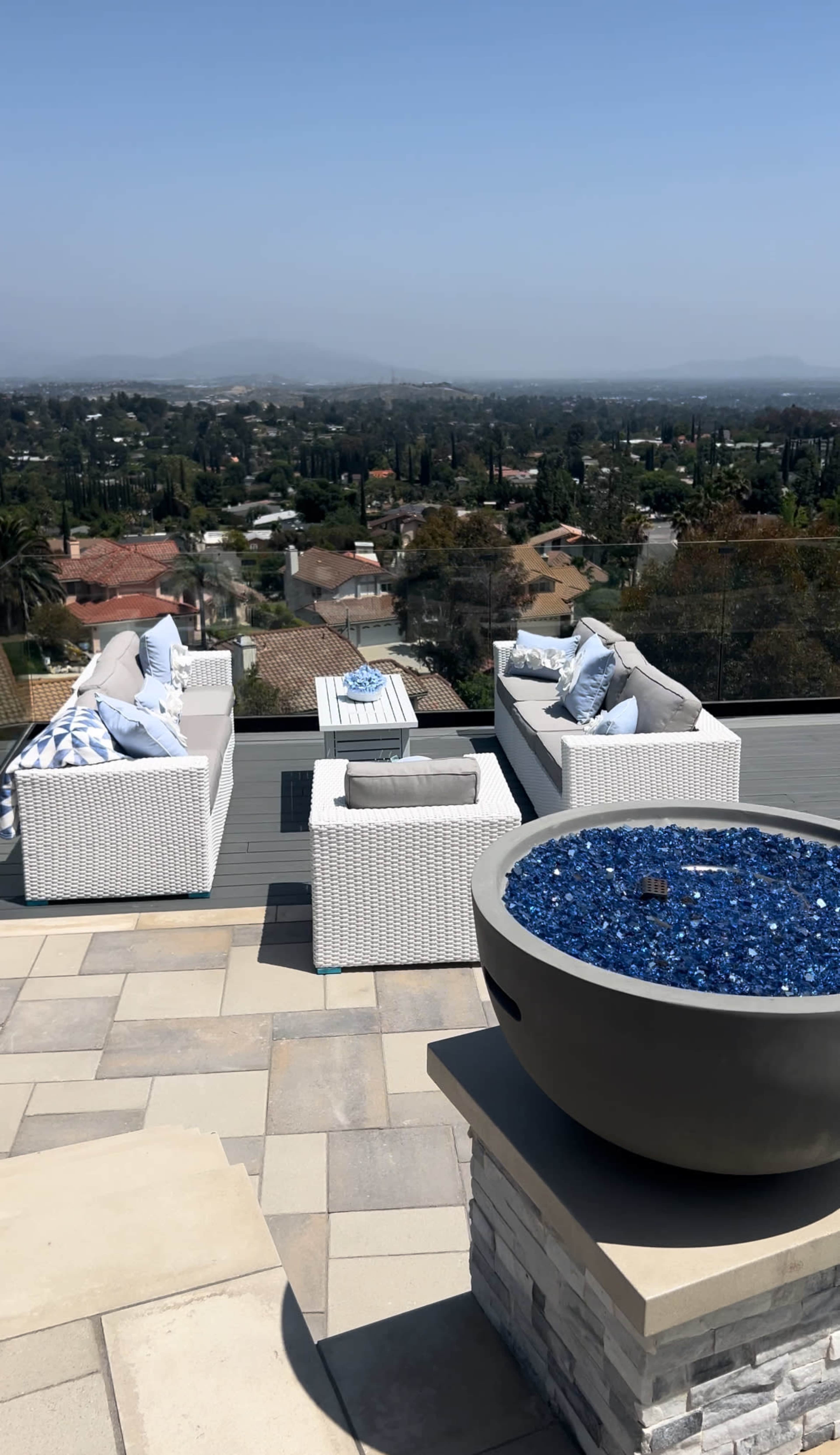 A rooftop patio with white wicker furniture arranged around a circular fire pit filled with blue glass stones, overlooking a landscaped view of homes and hills.