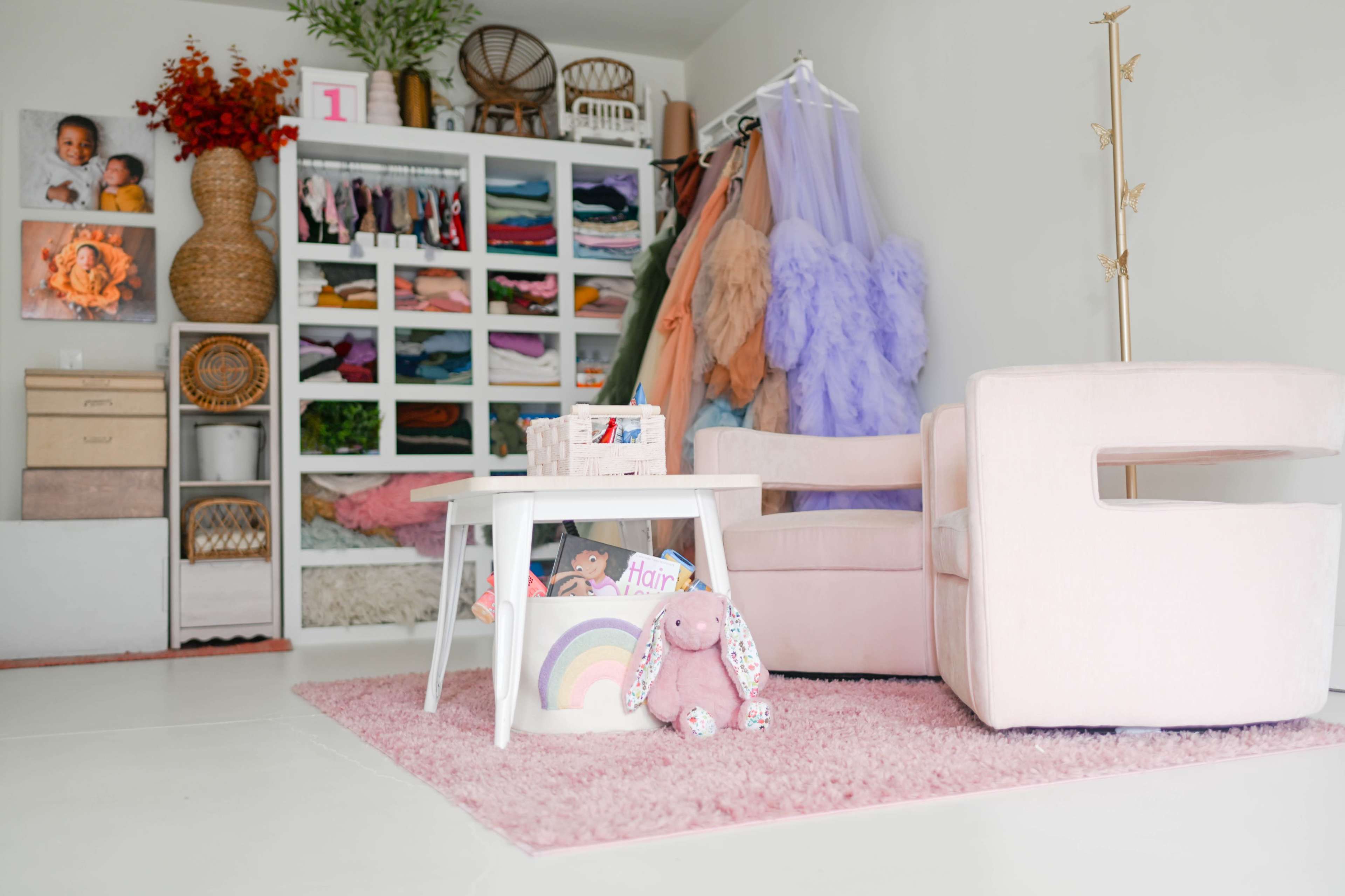 The image shows a well-organized room with a pink armchair, a small white table, and a shelf displaying colorful clothing and accessories for children, alongside a plush bunny and a rainbow decoration on the table.