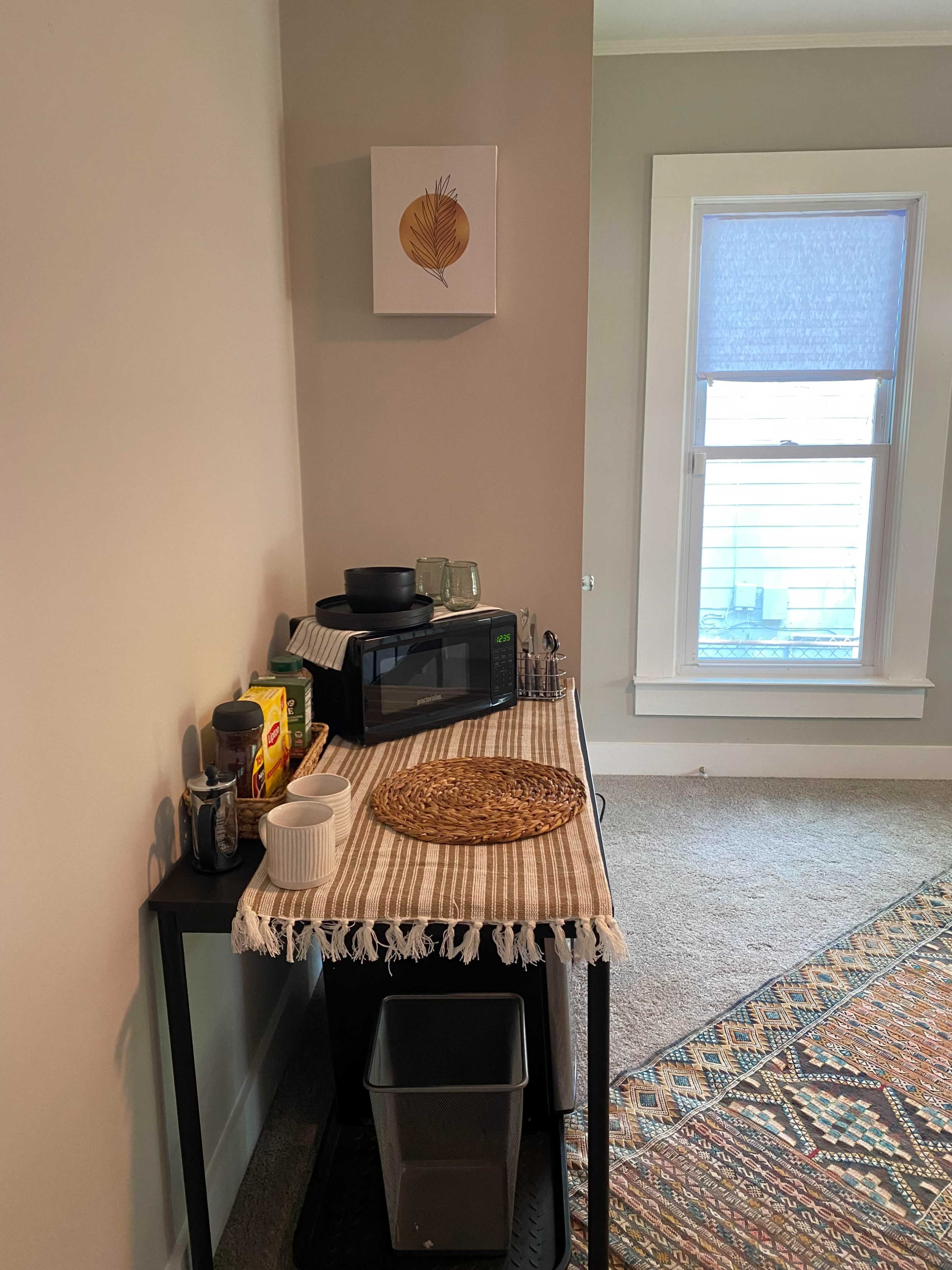 A small kitchen corner features a microwave, cooking utensils, a woven placemat, and a trash bin, with a window and wall art above.