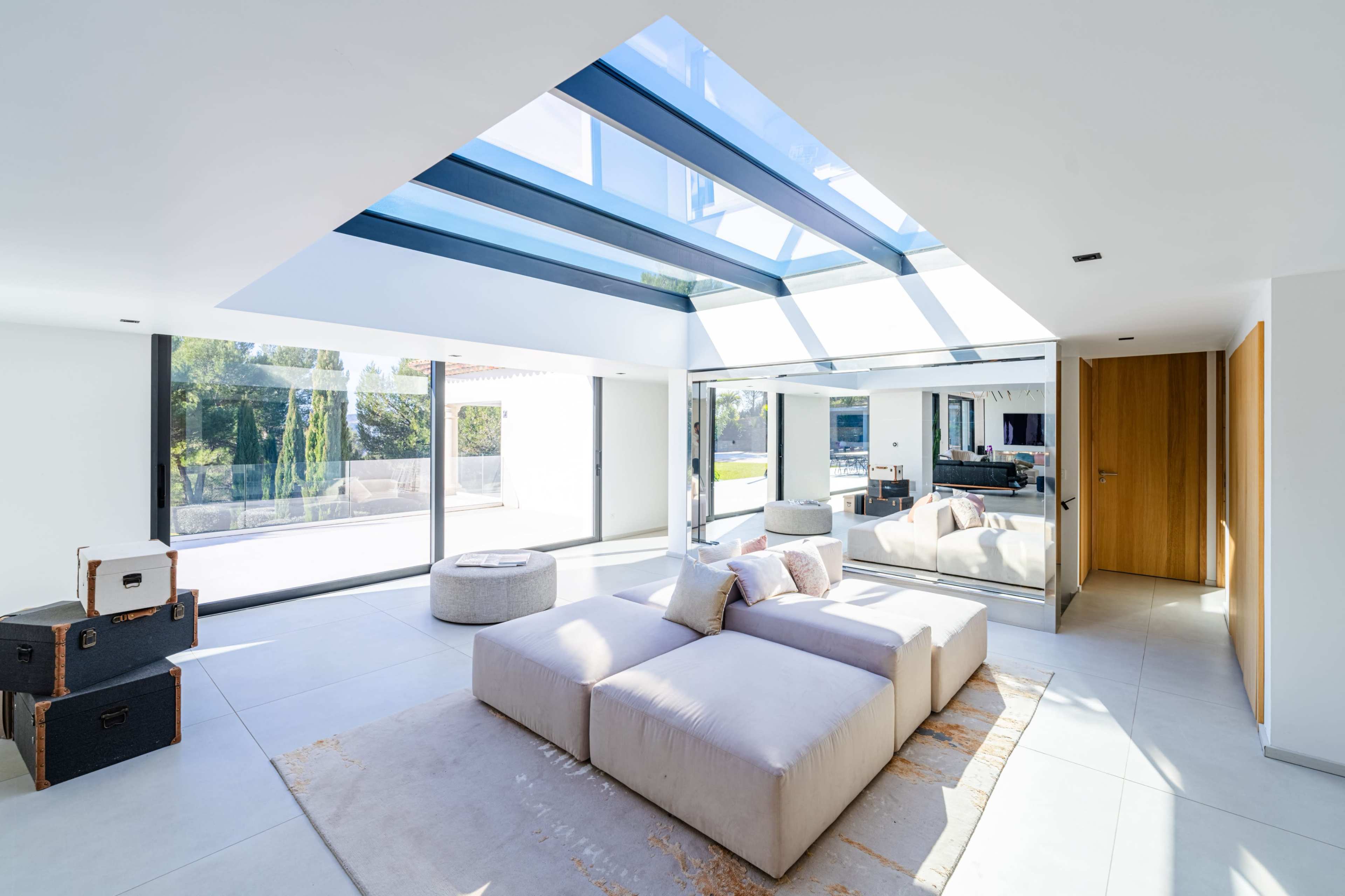 The image shows a modern living room featuring a sectional sofa, a round ottoman, large windows allowing natural light, and a skylight above.