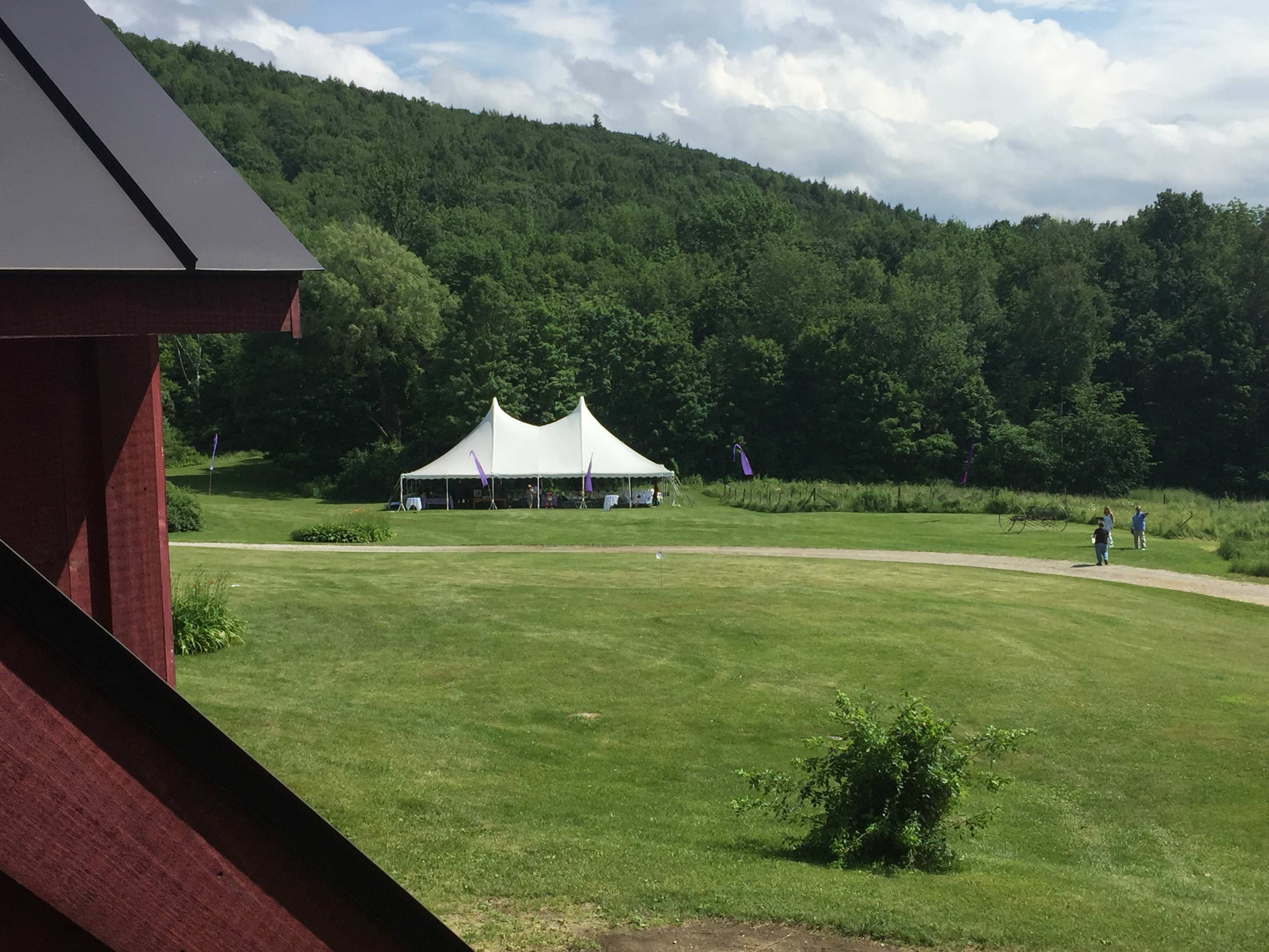 A large white tent is set up on a grassy area surrounded by trees and hills, with a few people walking nearby.