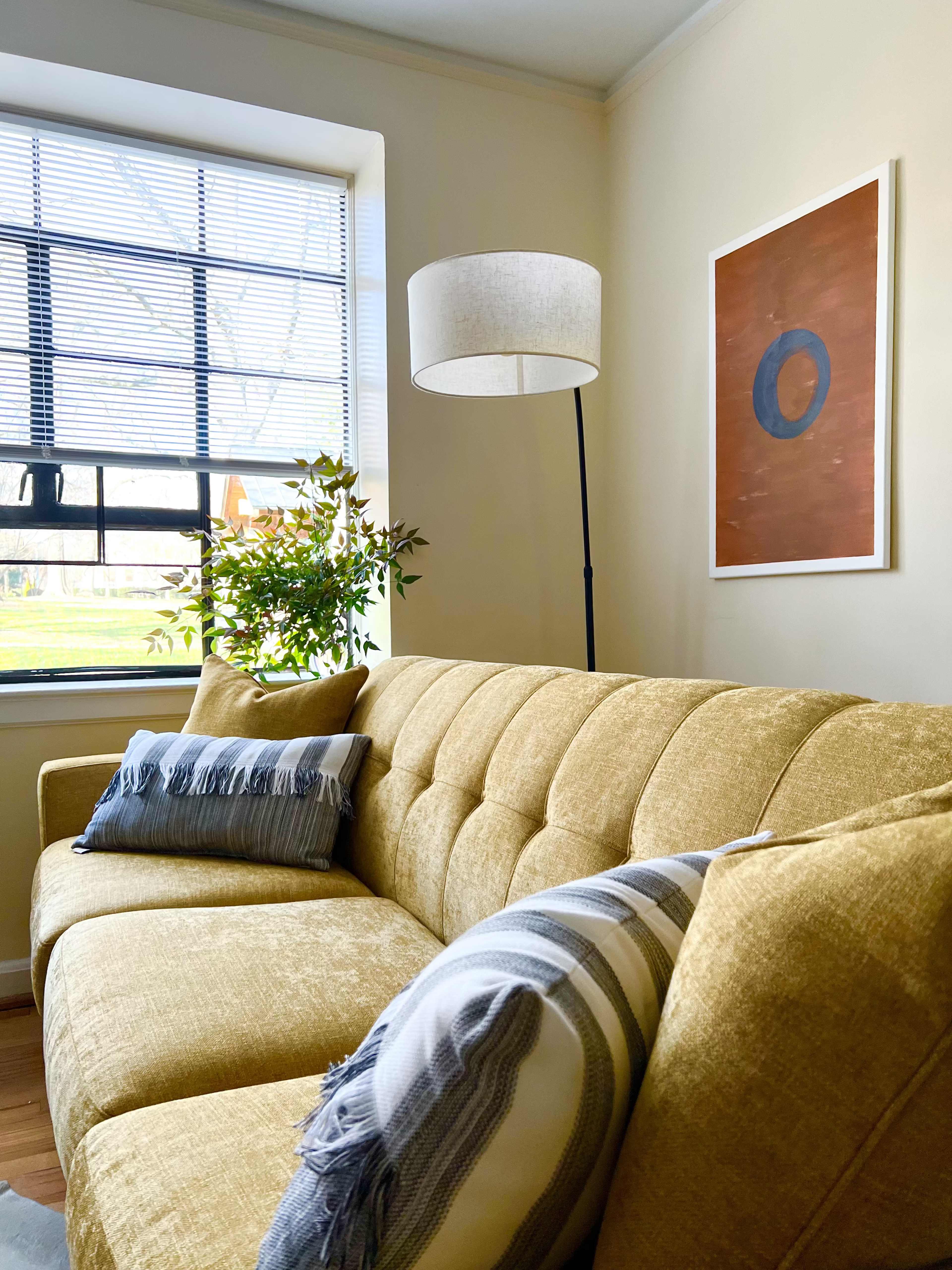 A yellow tufted sofa with decorative pillows sits under a window with blinds, beside a floor lamp and a framed artwork on the wall.