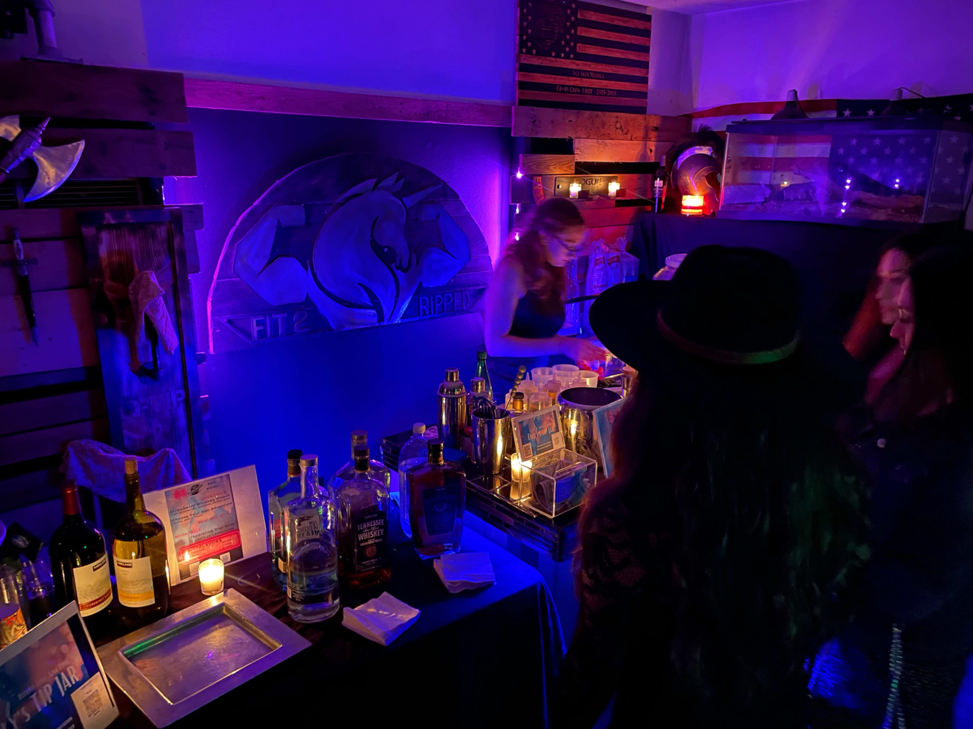 A dimly lit bar area decorated with purple lighting, featuring various bottles of alcohol and two women standing near the counter.